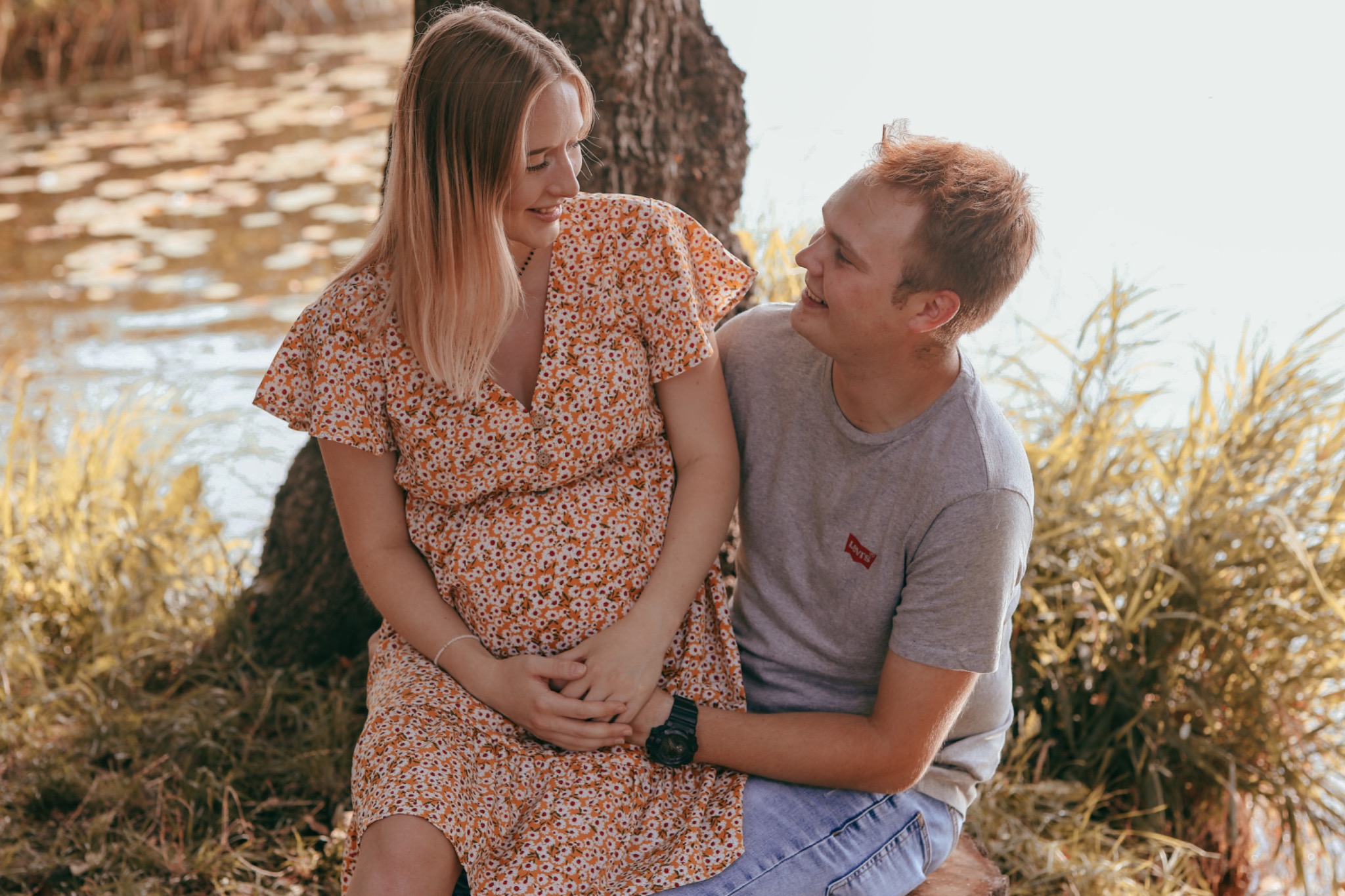 Uśmiechnięta kobieta w kwiecistej sukience i mężczyzna w szarej koszulce siedzą obok drzewa nad wodą, trzymając się za ręce.