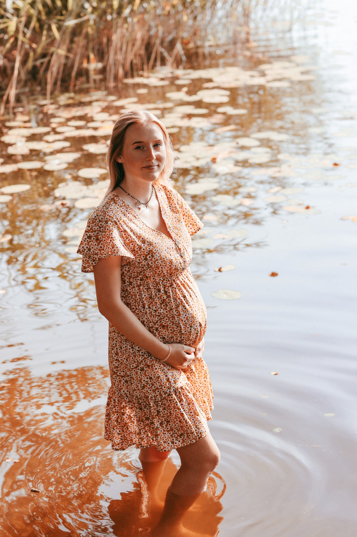 Kobieta w kwiecistej sukience ciążowej stoi w płytkiej wodzie jeziora, z liliami wodnymi w tle, trzymając ręce na brzuchu.