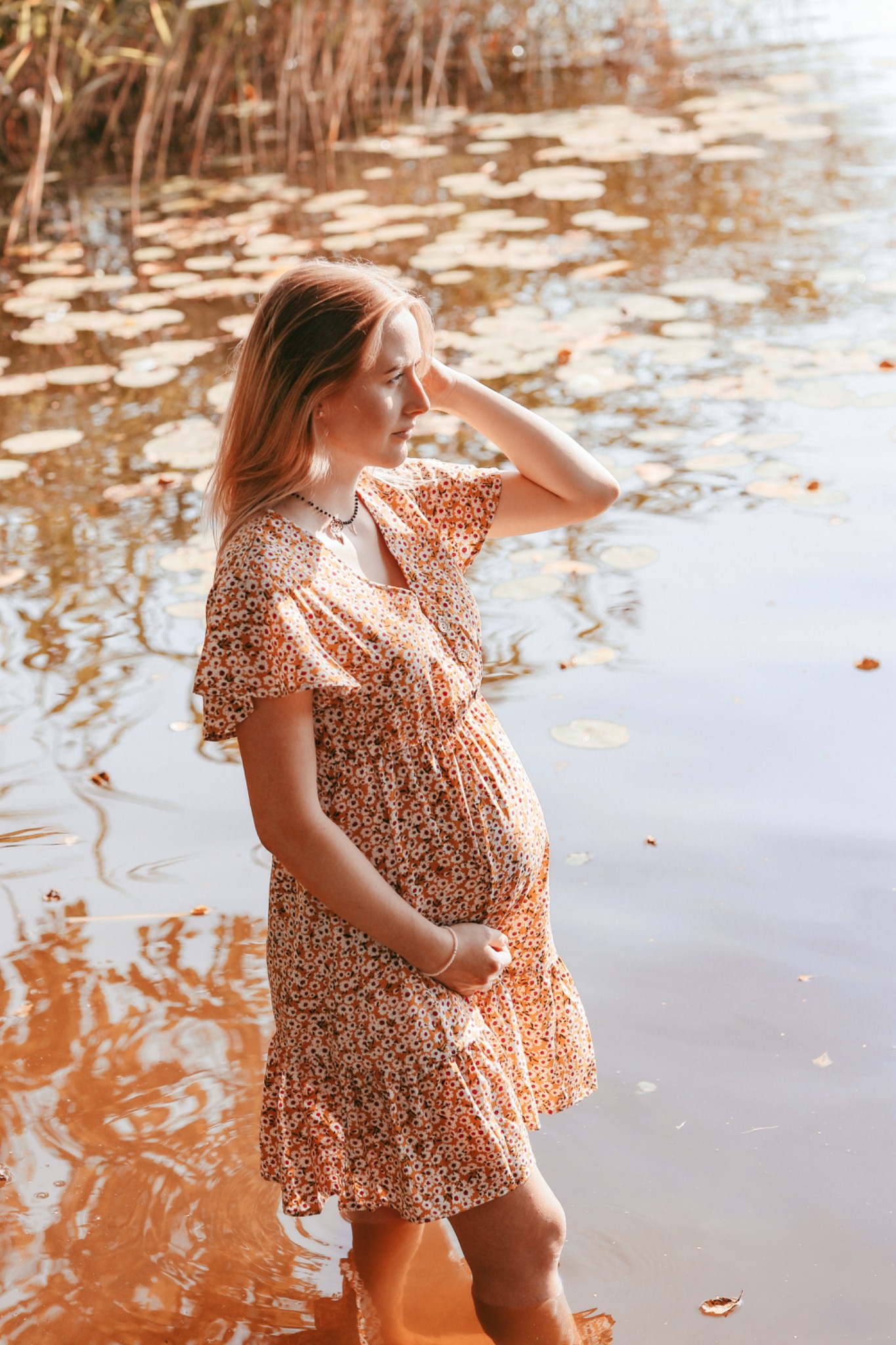 Kobieta w kwiecistej sukience ciążowej stoi w wodzie jeziora, z dłonią na brzuchu i drugą uniesioną do włosów, patrząc w dal na tle lilii wodnych i trzcin.