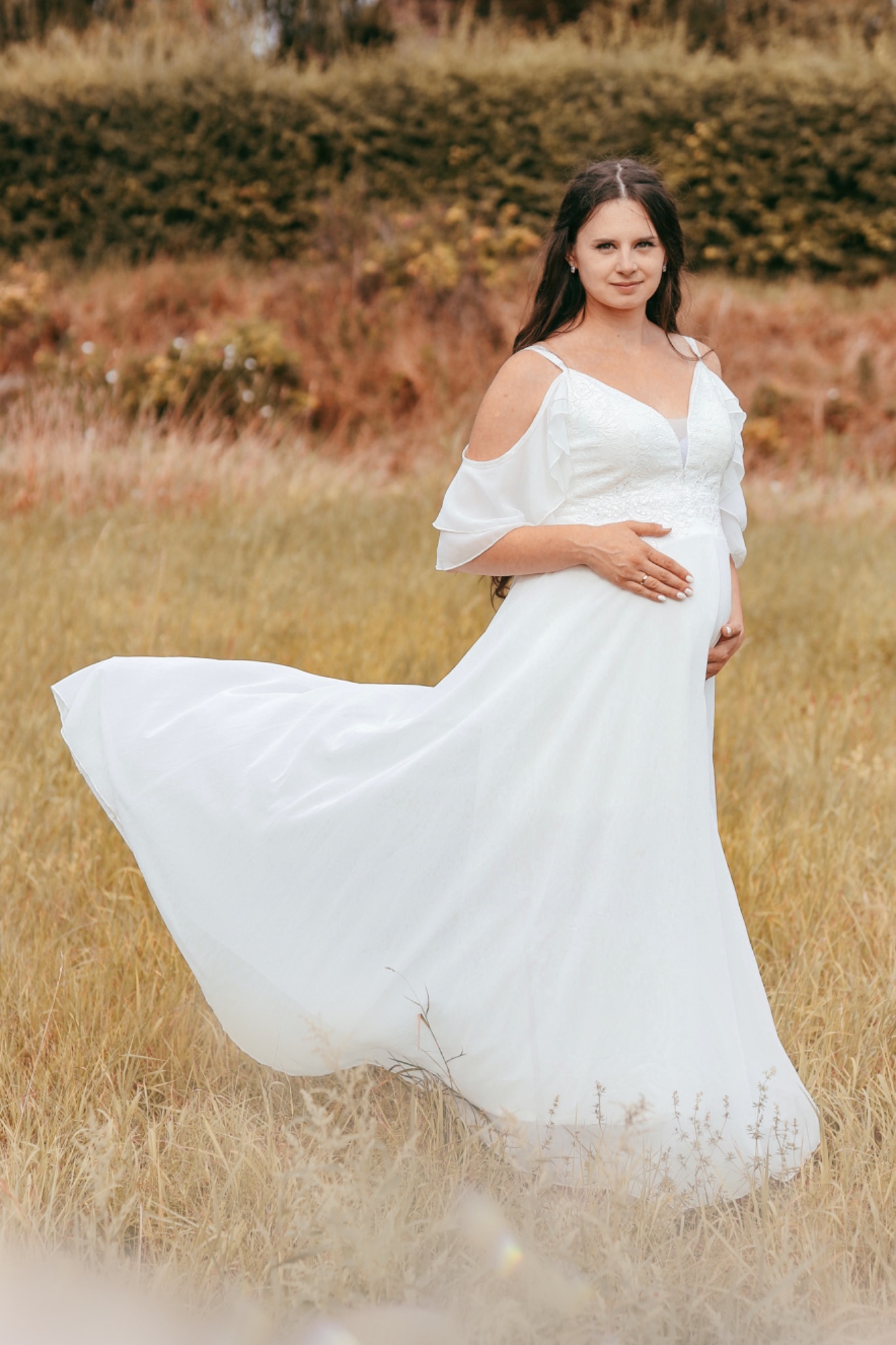 Kobieta w zwiewnej białej sukni ciążowej pozuje na łące w Szczecinku, dłoń spoczywa na brzuchu, delikatny uśmiech.