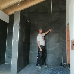 Usługi glazurnicze i kompleksowe wykończenia wnętrz - Mężczyzna w czapce mierzy wysokość ściany wyłożonej ciemnymi, kwadratowymi płytkami w łazience w trakcie remontu. Widoczne drewniane belki stropowe i resztki zaprawy na podłodze.