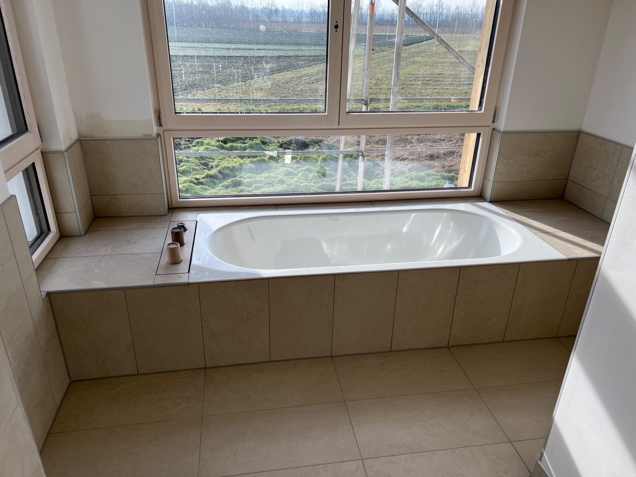Wanna obudowana beżowymi płytkami w łazience z oknem na pole, widoczne akcesoria łazienkowe.