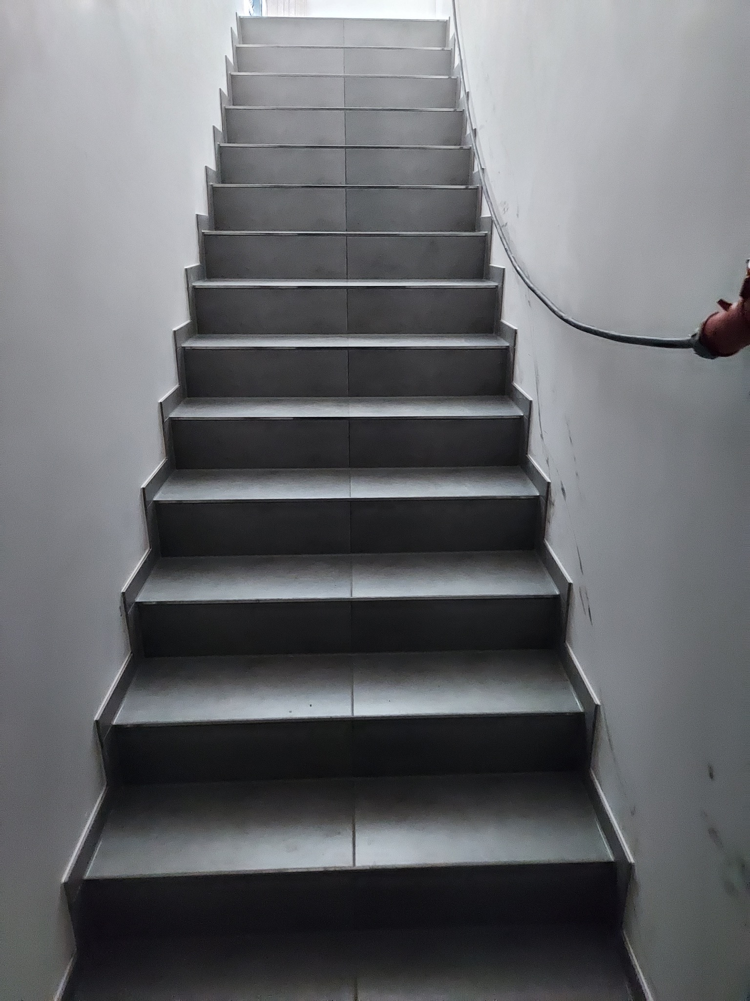 Schody wyłożone szarymi płytkami ceramicznymi, widok z dołu, z widoczną instalacją przeciwpożarową na ścianie.