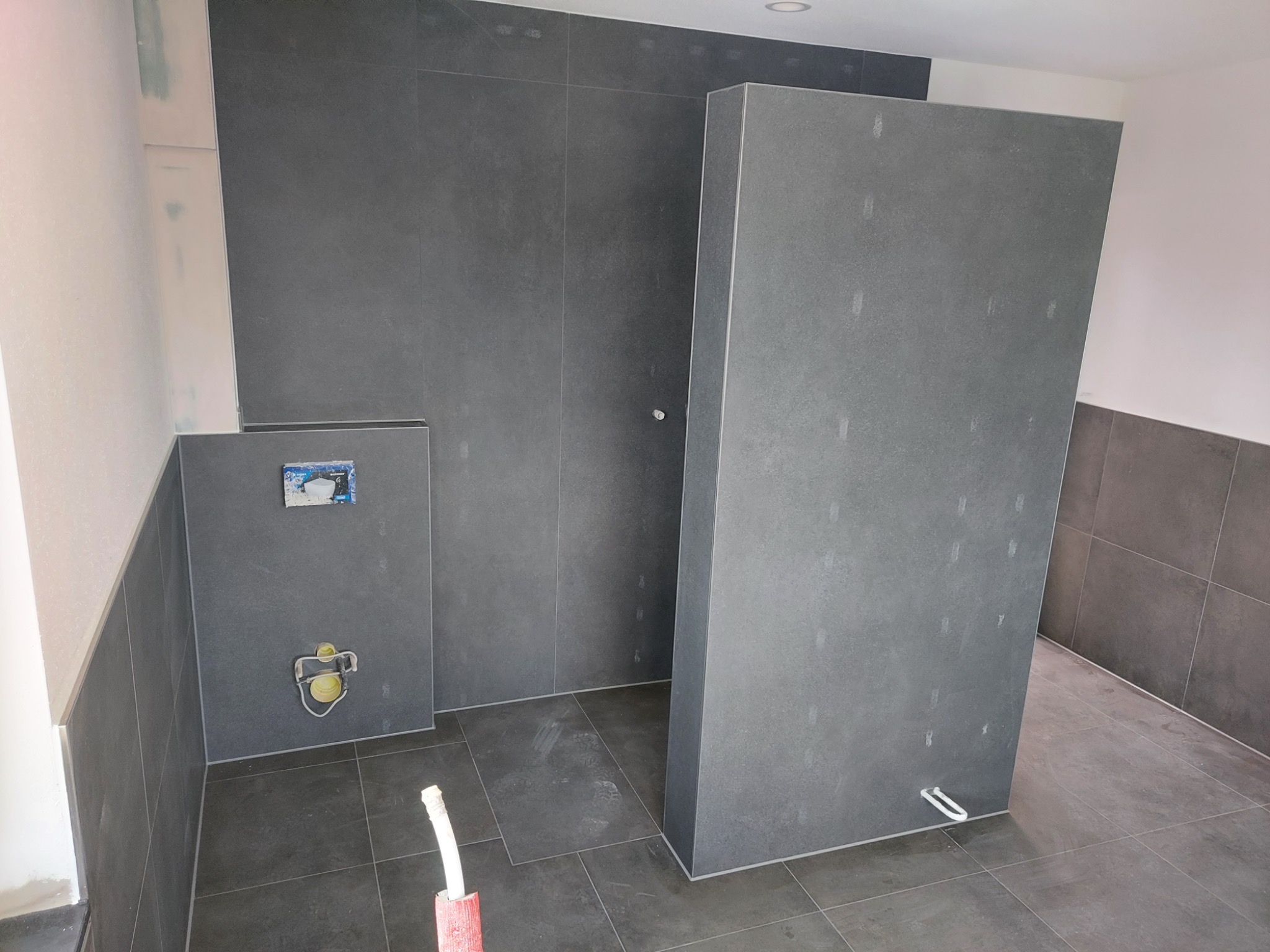 Wnętrze łazienki w trakcie remontu, z szarymi płytkami na ścianach i podłodze, widoczne instalacje hydrauliczne i fragment narzędzia malarskiego w rogu.