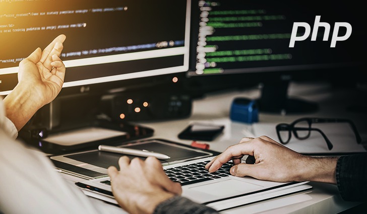 Programista pracuje przy biurku z dwoma monitorami, na których wyświetlany jest kod. Na pierwszym planie widoczne dłonie piszące na laptopie, a obok tablet graficzny z rysikiem. Na jednym...