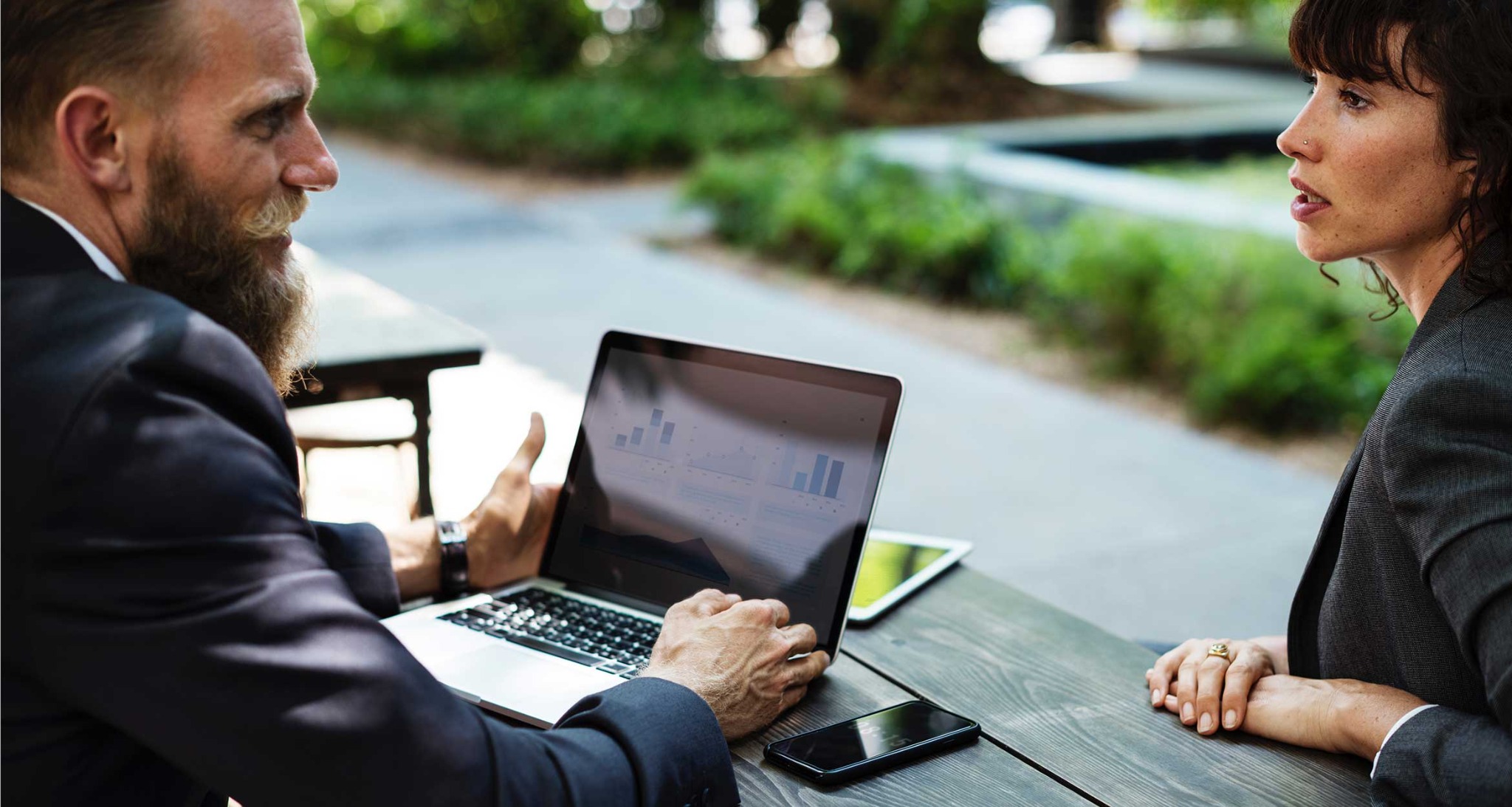 Mężczyzna z brodą w garniturze prezentuje na laptopie wykresy słupkowe kobiecie z kolczykiem w nosie, siedząc przy drewnianym stole na zewnątrz.