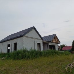 Mp-bud - Dwa budynki w stanie surowym otwartym z częściowo położonym ciemnym pokryciem na dachach. Na jednym budynku widoczna więźba dachowa. Na drugim planie inne budynki i zieleń.