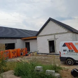 Dom w stanie surowym otwartym z częściowo położonym ciemnoszarym pokryciem dachowym, widoczny samochód dostawczy z logo firmy dekarskiej i stertą materiałów budowlanych przed budynkiem.