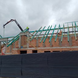 Mp-bud - Budowa dachu z drewnianą konstrukcją i dźwigiem na tle pochmurnego nieba, widok zza czarnego ogrodzenia. Mur z czerwonej cegły w trakcie wznoszenia.