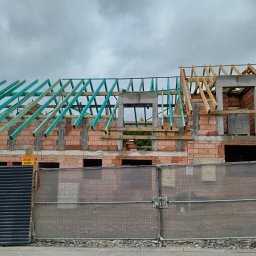 Mp-bud - Budowa domu w trakcie wznoszenia więźby dachowej, z widocznymi elementami drewnianymi w kolorze naturalnym i zielonym, na tle pochmurnego nieba.