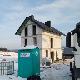 Mp-bud - Surowy dom z czarnym dachem w zimowym krajobrazie, widoczna przenośna toaleta i elementy budowlane. Dom w trakcie budowy, z widoczną konstrukcją dachu.