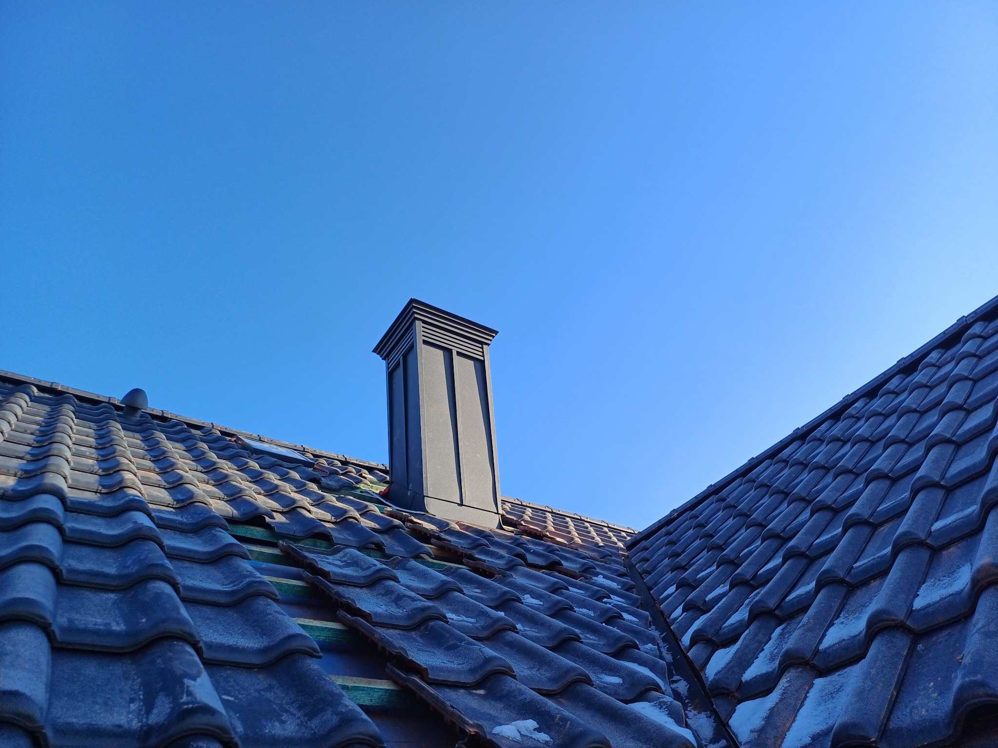 Ciemna dachówka ceramiczna z kominem na tle błękitnego nieba, widoczne fragmenty łat pod dachówką.