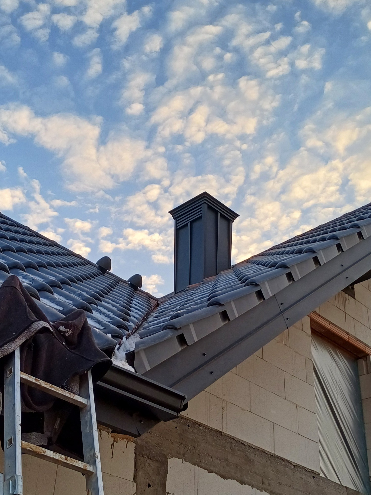 Nowy, grafitowy dach z ceramicznej dachówki karpiówki, widok z dołu na komin i obróbki blacharskie, fragment drabiny oparty o rynnę, budynek w trakcie wykończenia.