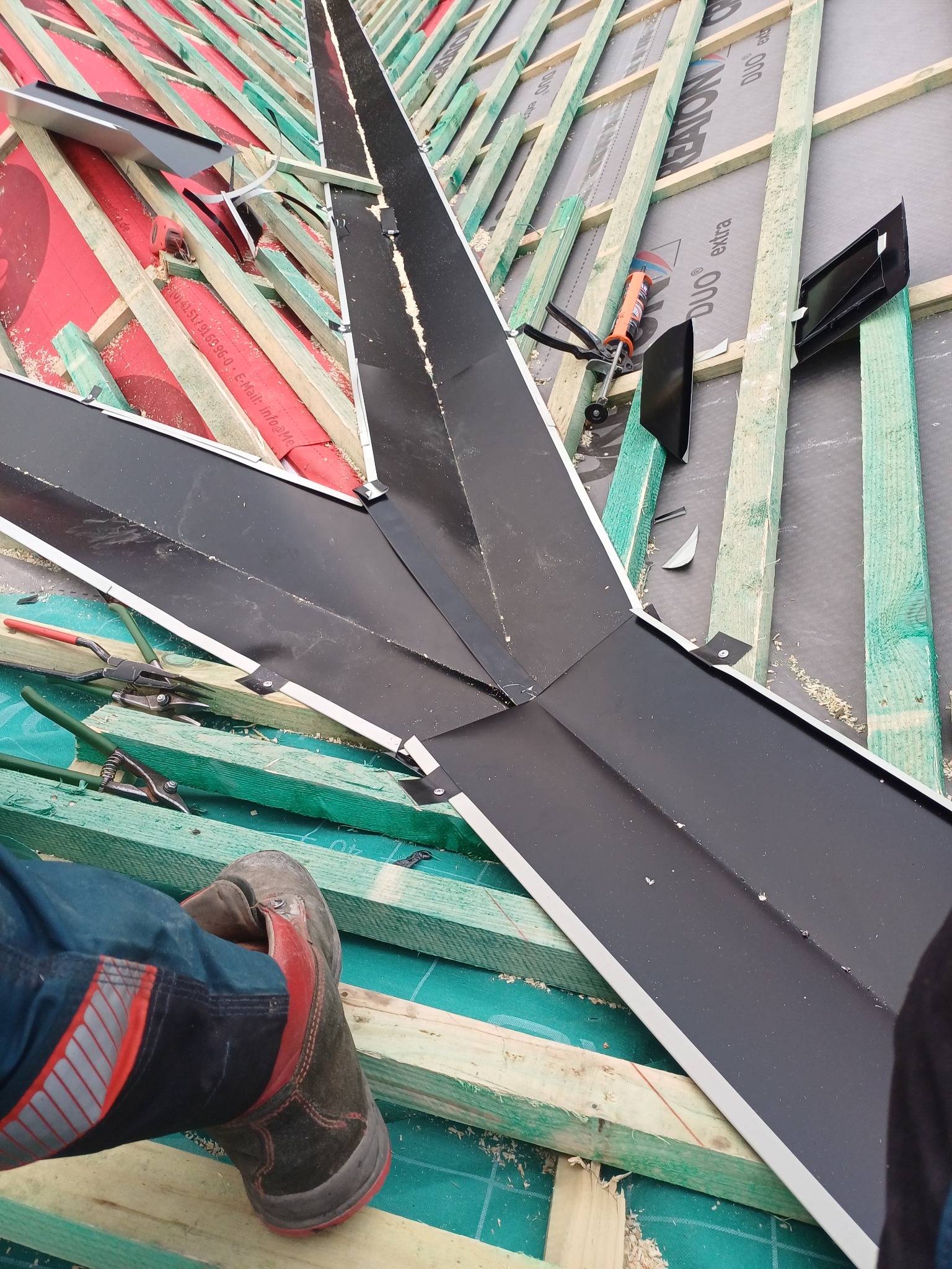 Montaż obróbki blacharskiej na dachu z widoczną więźbą, narzędziami i fragmentem buta pracownika.