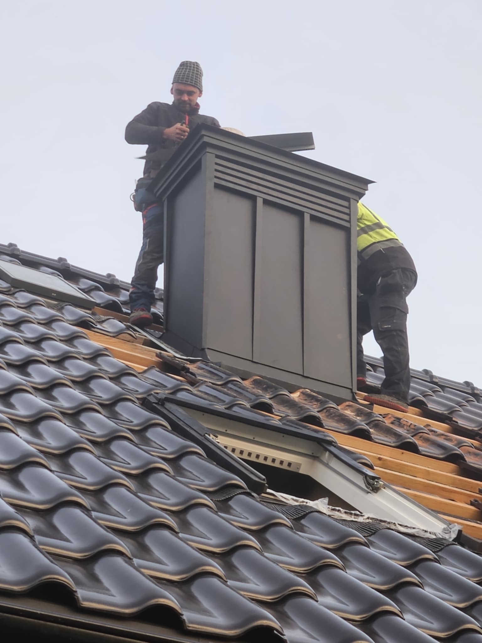Dwóch dekarzy montuje ciemnoszary komin wentylacyjny na dachu z ciemnej dachówki ceramicznej, widoczne elementy drewniane konstrukcji dachu i okno dachowe.
