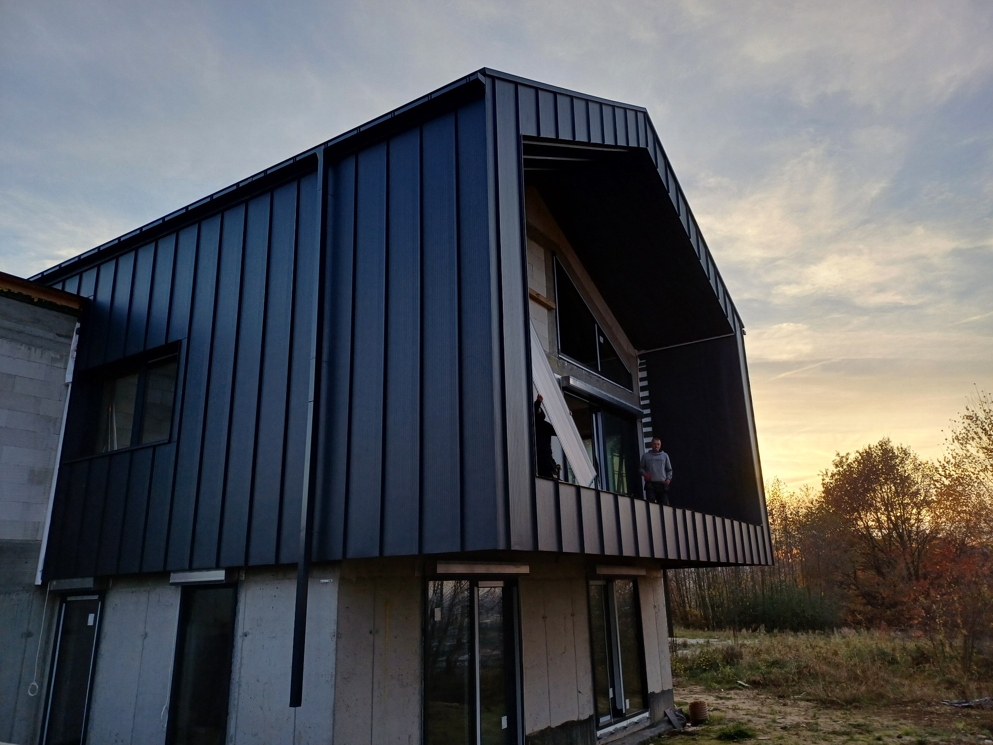 Nowoczesny dom w trakcie budowy z ciemną, pionową okładziną elewacyjną i dachem z blachy na tle zachodzącego słońca. Widoczny pracownik na balkonie.