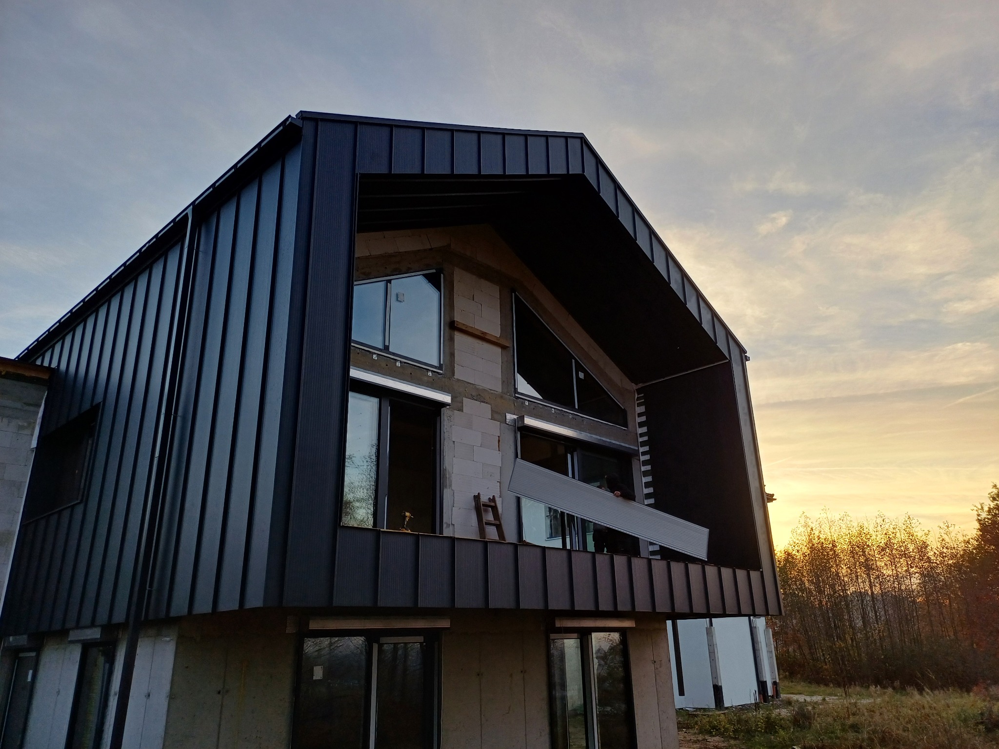 Nowoczesny dom w trakcie wykańczania, widoczna czarna blacha na dachu i elewacji, okna jeszcze bez wykończenia, w tle drzewa i zachodzące słońce.