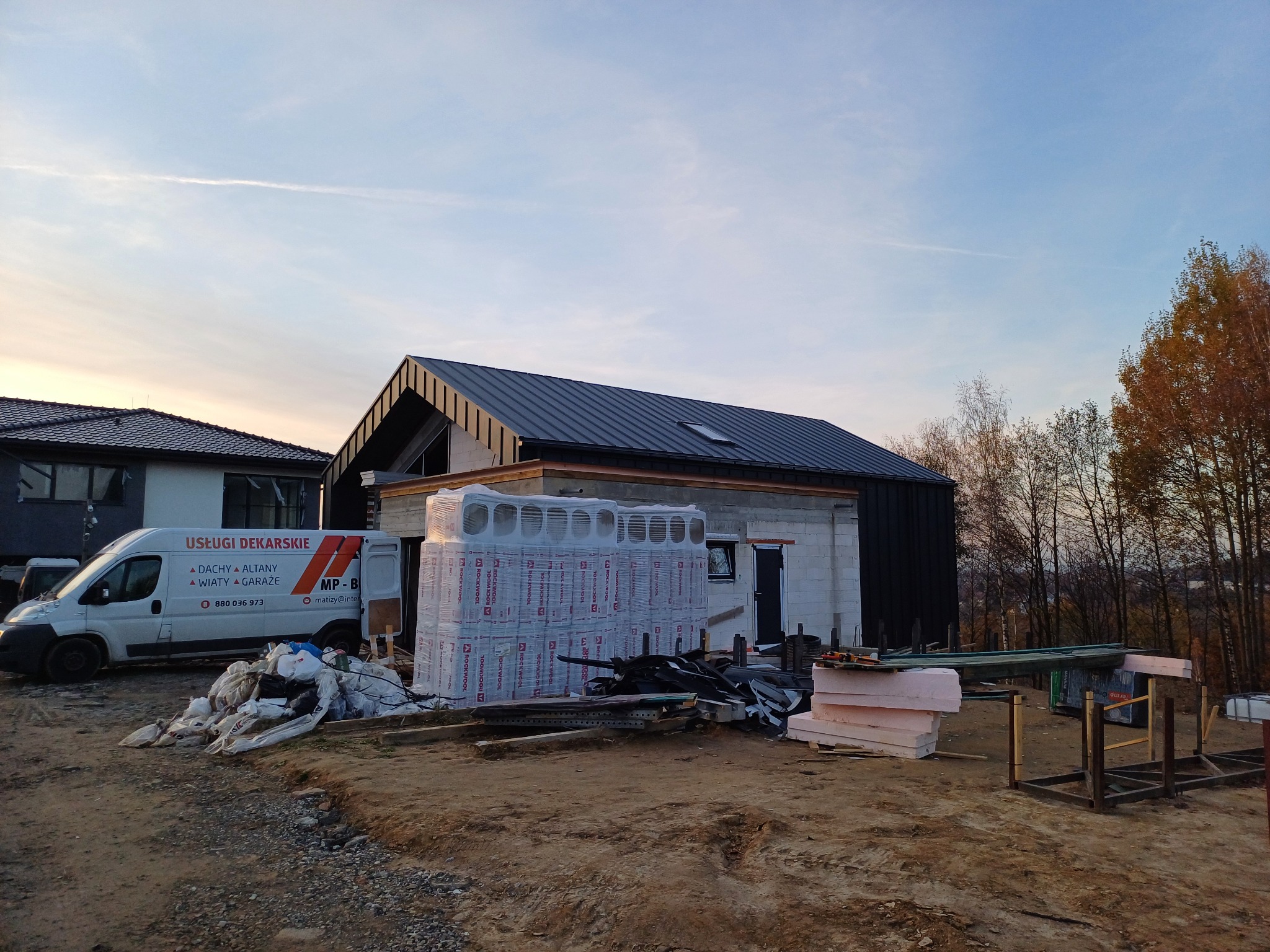 Nowo wybudowany dom z czarnym, metalowym pokryciem dachowym, widoczny z poziomu gruntu, z materiałami budowlanymi i firmowym samochodem dostawczym na pierwszym planie.