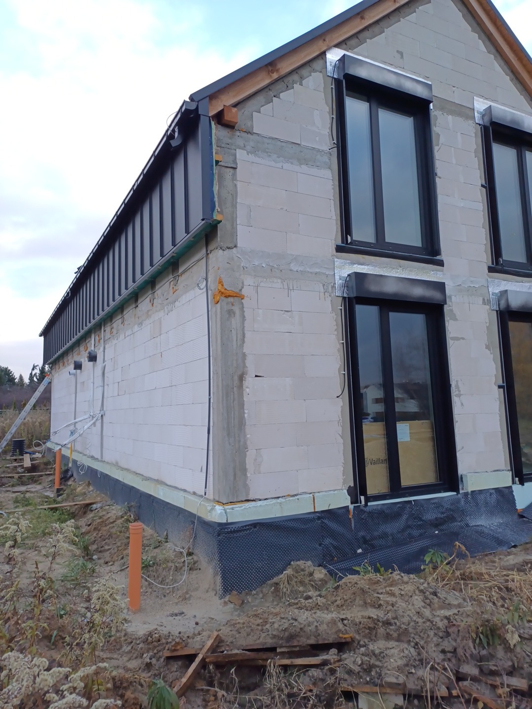 Niedokończony budynek z zamontowanymi oknami i czarnymi rynnami, widoczne elementy konstrukcji dachu i izolacji fundamentów.