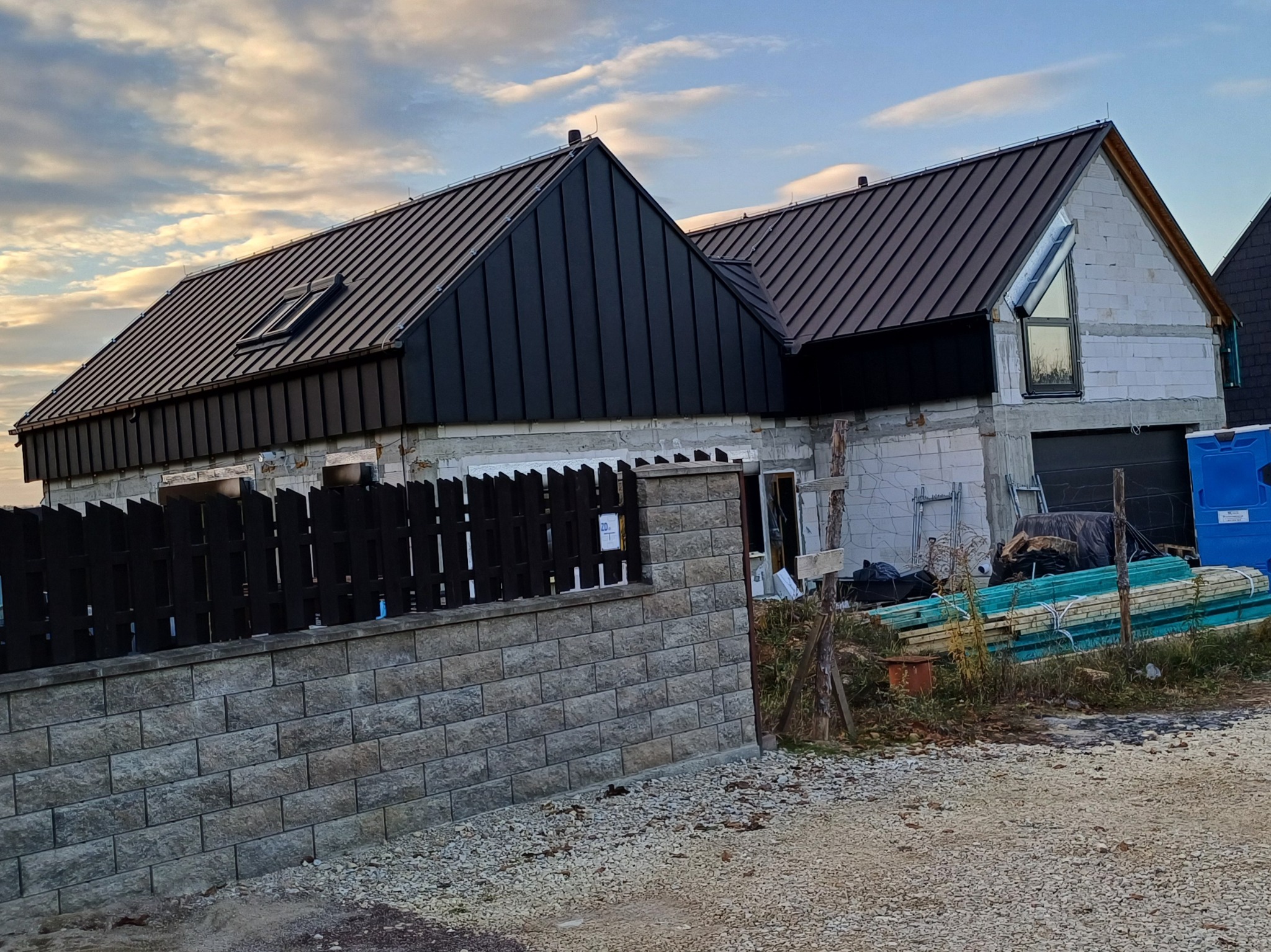 Dom w stanie surowym zamkniętym z ciemną blachodachówką, widoczne materiały budowlane i ogrodzenie z betonowych bloczków.