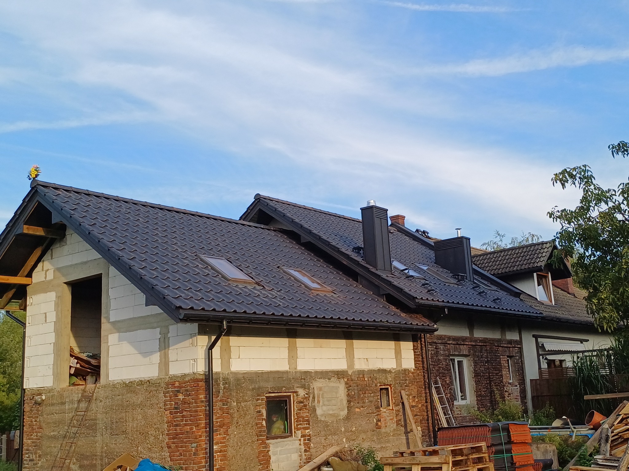 Dom w trakcie remontu dachu z widocznymi oknami dachowymi, nową rynną i ozdobą na szczycie, z elementami starej i nowej konstrukcji.