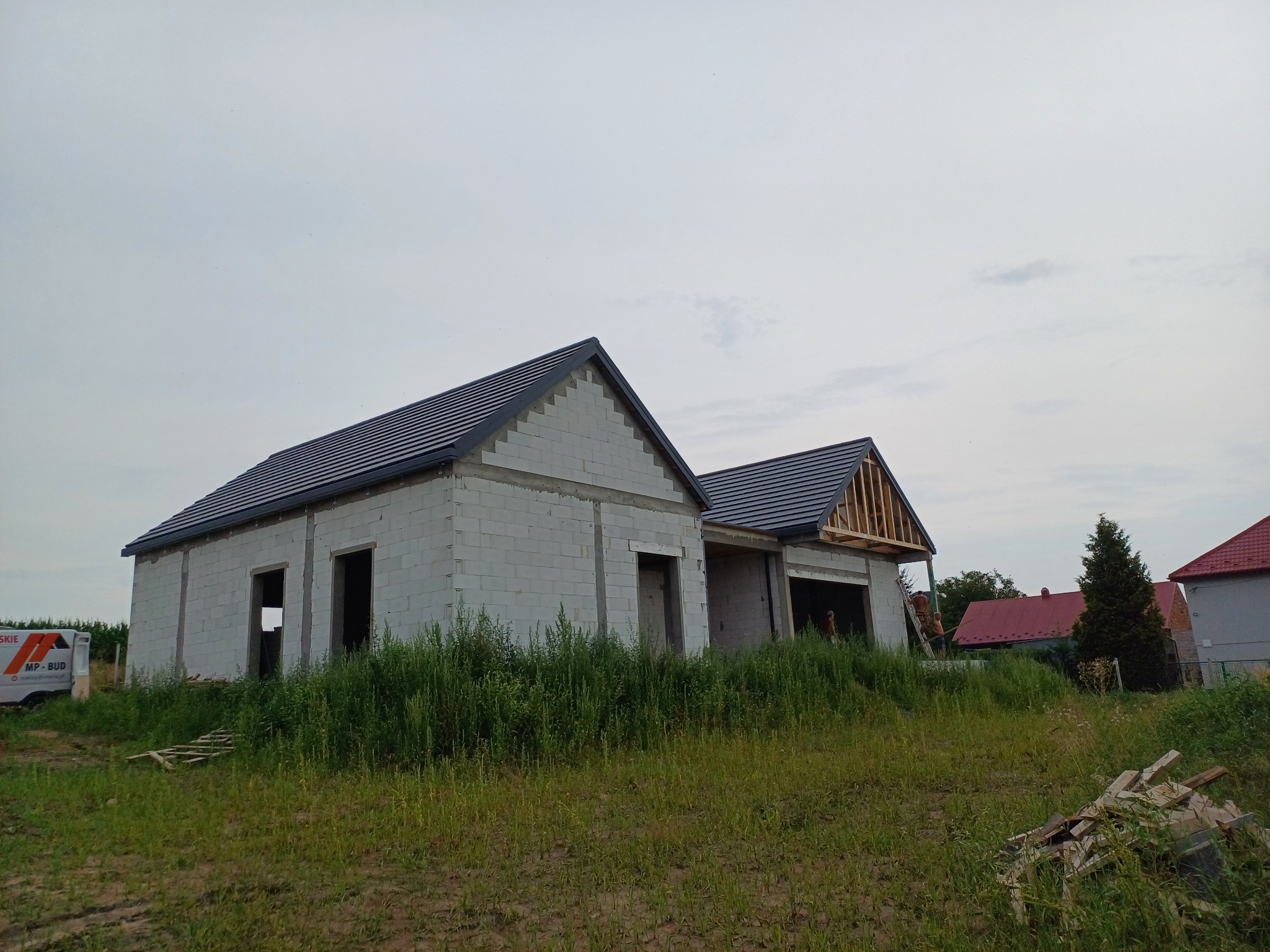 Dwa budynki w stanie surowym otwartym z częściowo położonym ciemnym pokryciem na dachach. Na jednym budynku widoczna więźba dachowa. Na drugim planie inne budynki i zieleń.