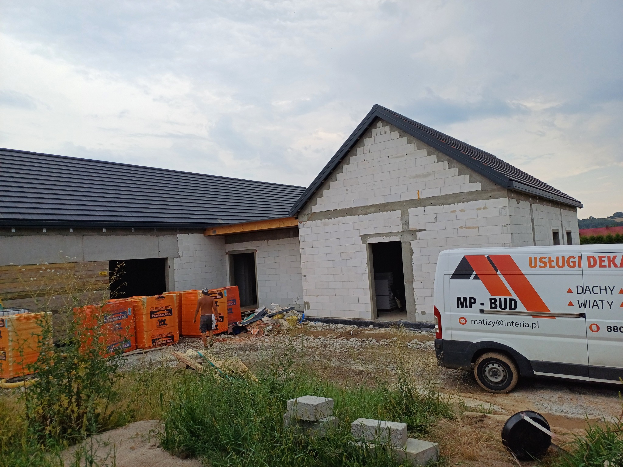Dom w stanie surowym otwartym z częściowo położonym ciemnoszarym pokryciem dachowym, widoczny samochód dostawczy z logo firmy dekarskiej i stertą materiałów budowlanych przed budynkiem.