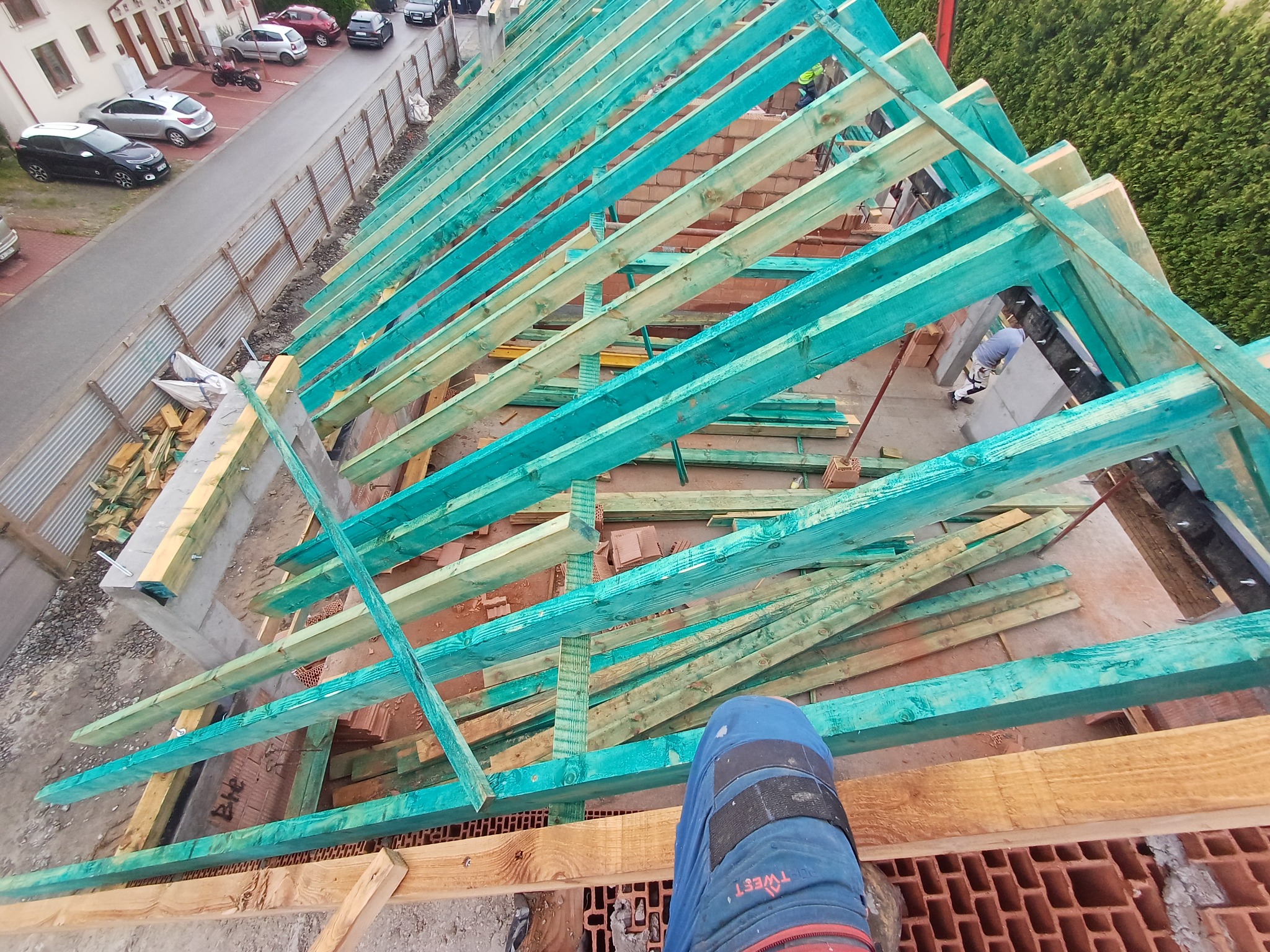 Wysoki kąt: drewniana, impregnowana na zielono więźba dachowa w trakcie montażu, widok z góry na konstrukcję i fragment nogi pracownika w niebieskich spodniach.