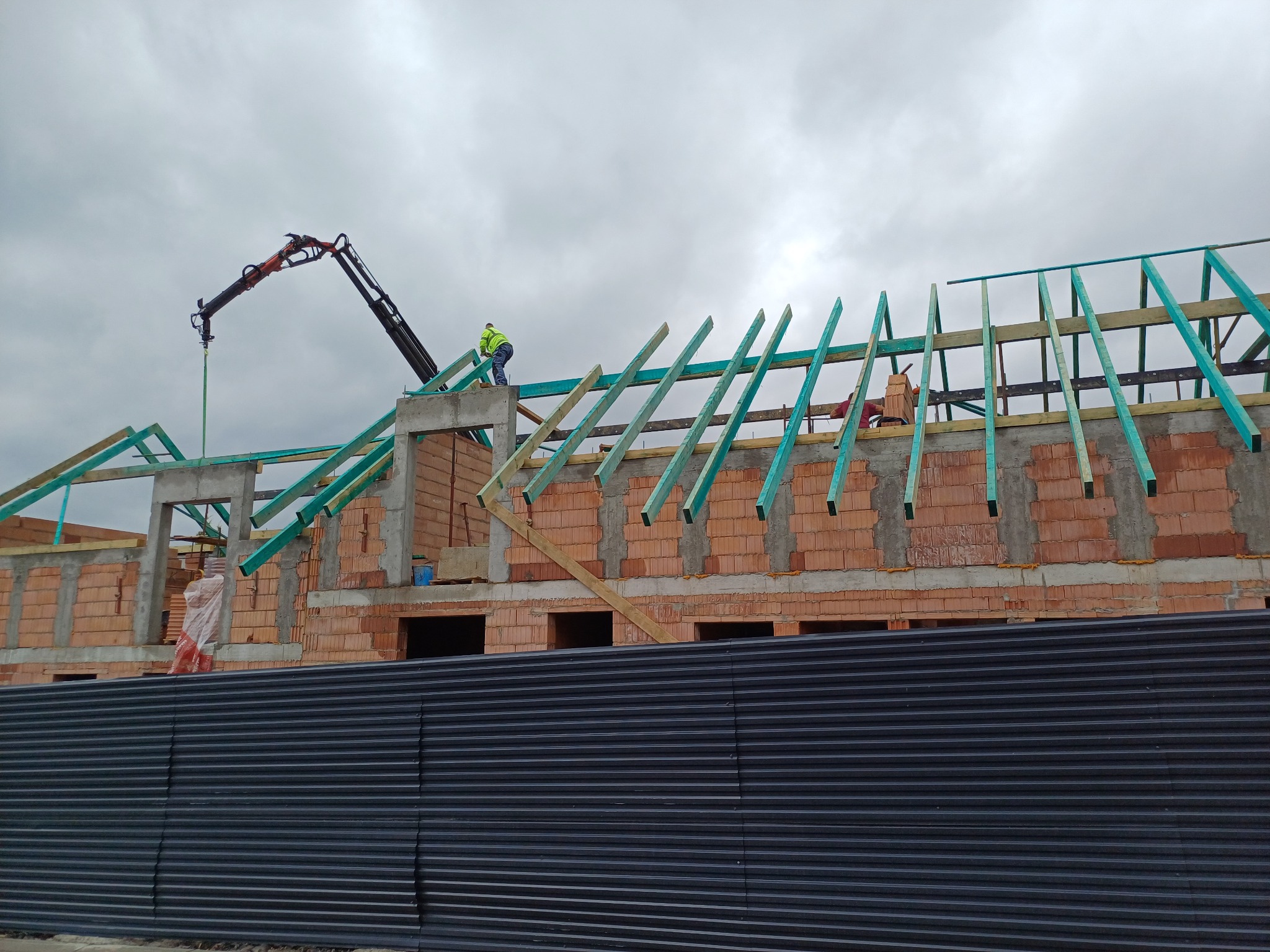 Budowa dachu z drewnianą konstrukcją i dźwigiem na tle pochmurnego nieba, widok zza czarnego ogrodzenia. Mur z czerwonej cegły w trakcie wznoszenia.