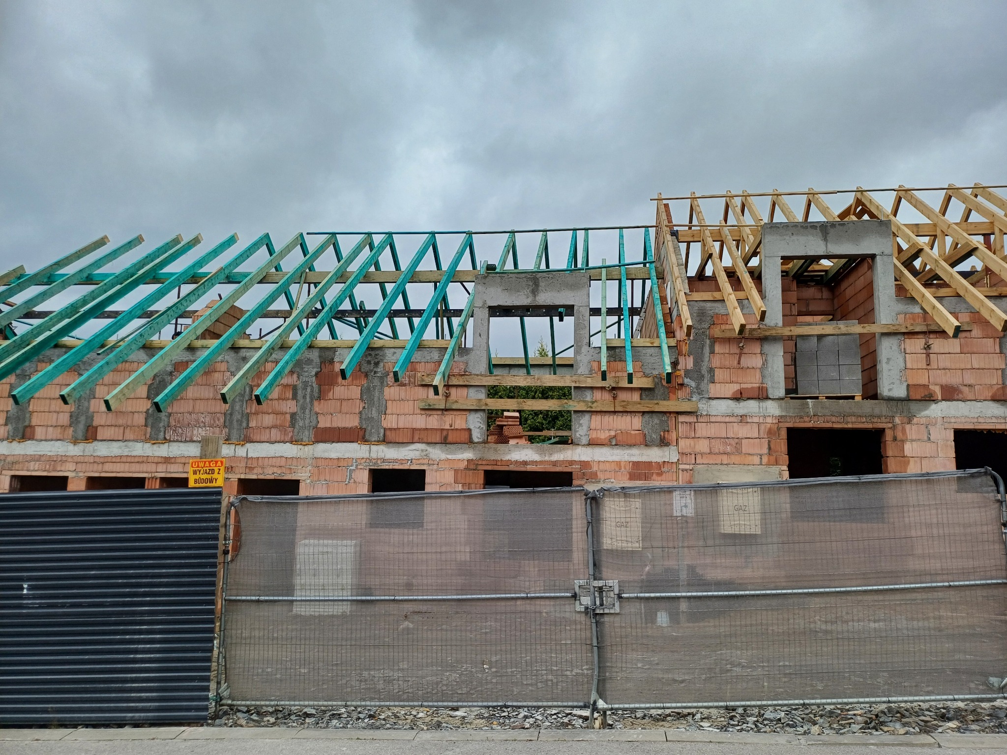 Budowa domu w trakcie wznoszenia więźby dachowej, z widocznymi elementami drewnianymi w kolorze naturalnym i zielonym, na tle pochmurnego nieba.