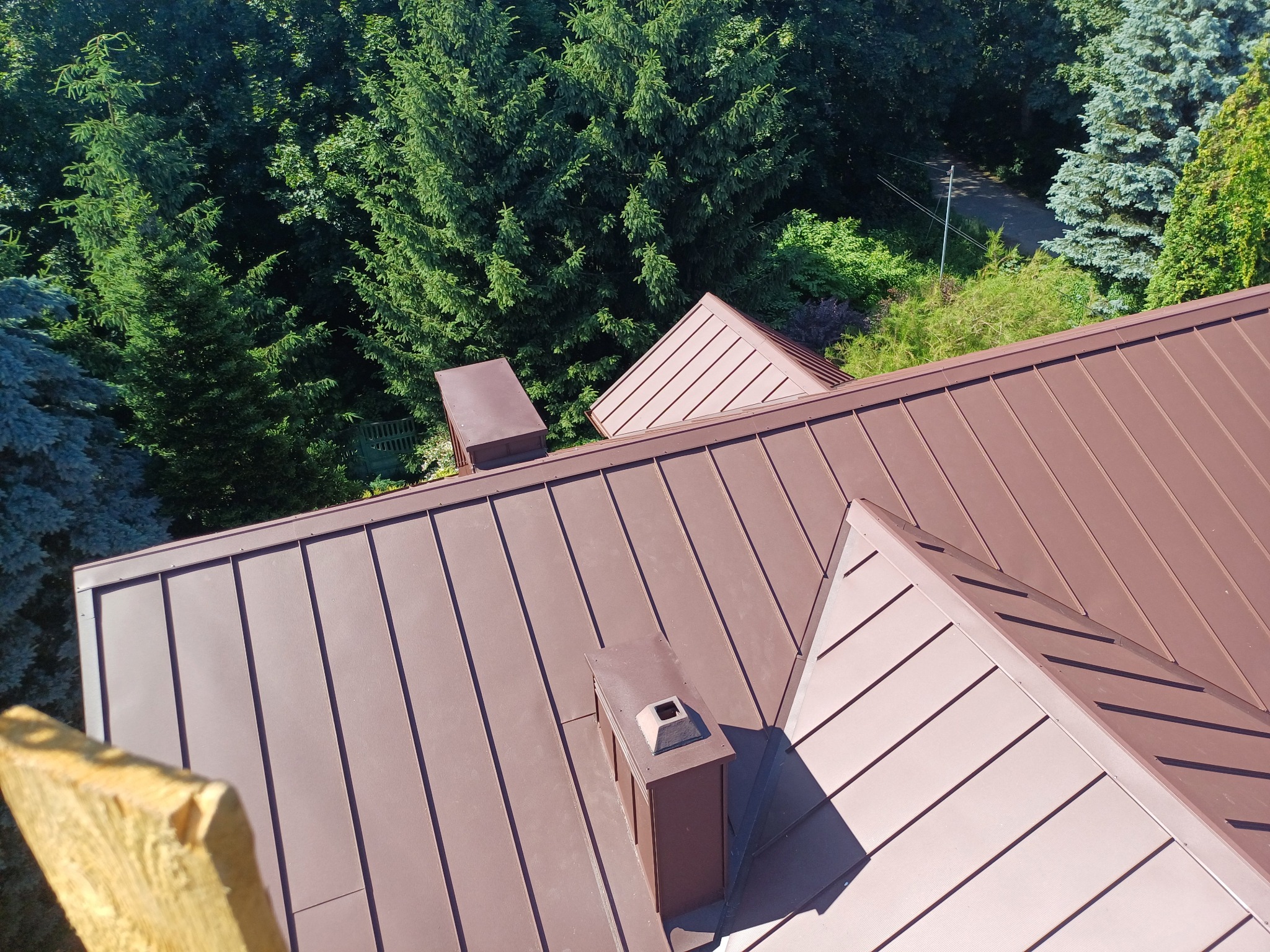 Widok z góry na brązowy dach z blachy trapezowej, kominy wentylacyjne i zielone drzewa w tle. Fragment deski w lewym dolnym rogu kadru.