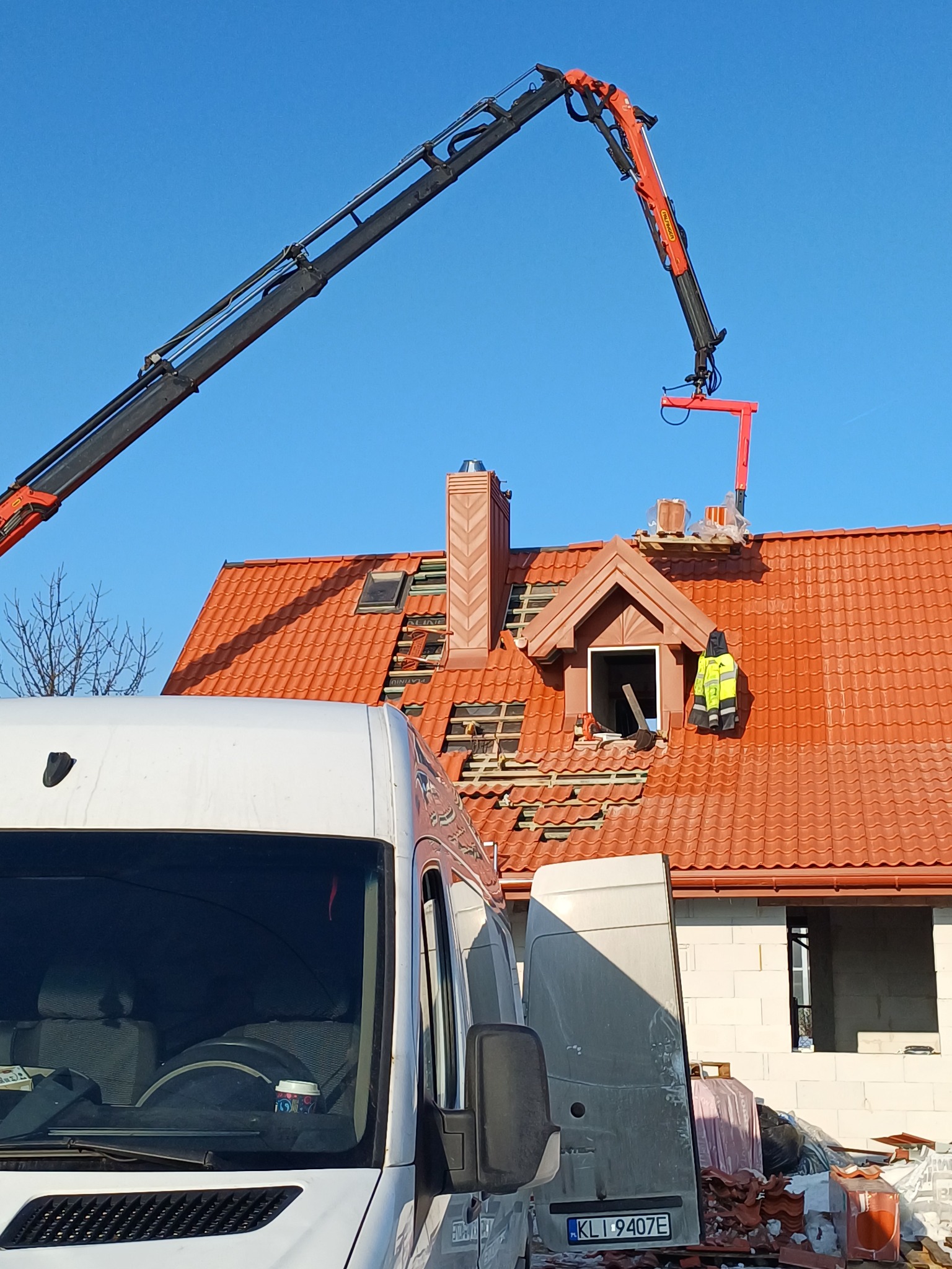 Czerwony dach w trakcie naprawy z użyciem dźwigu, widoczny pracownik w odblaskowej kamizelce. Na pierwszym planie biały samochód dostawczy.