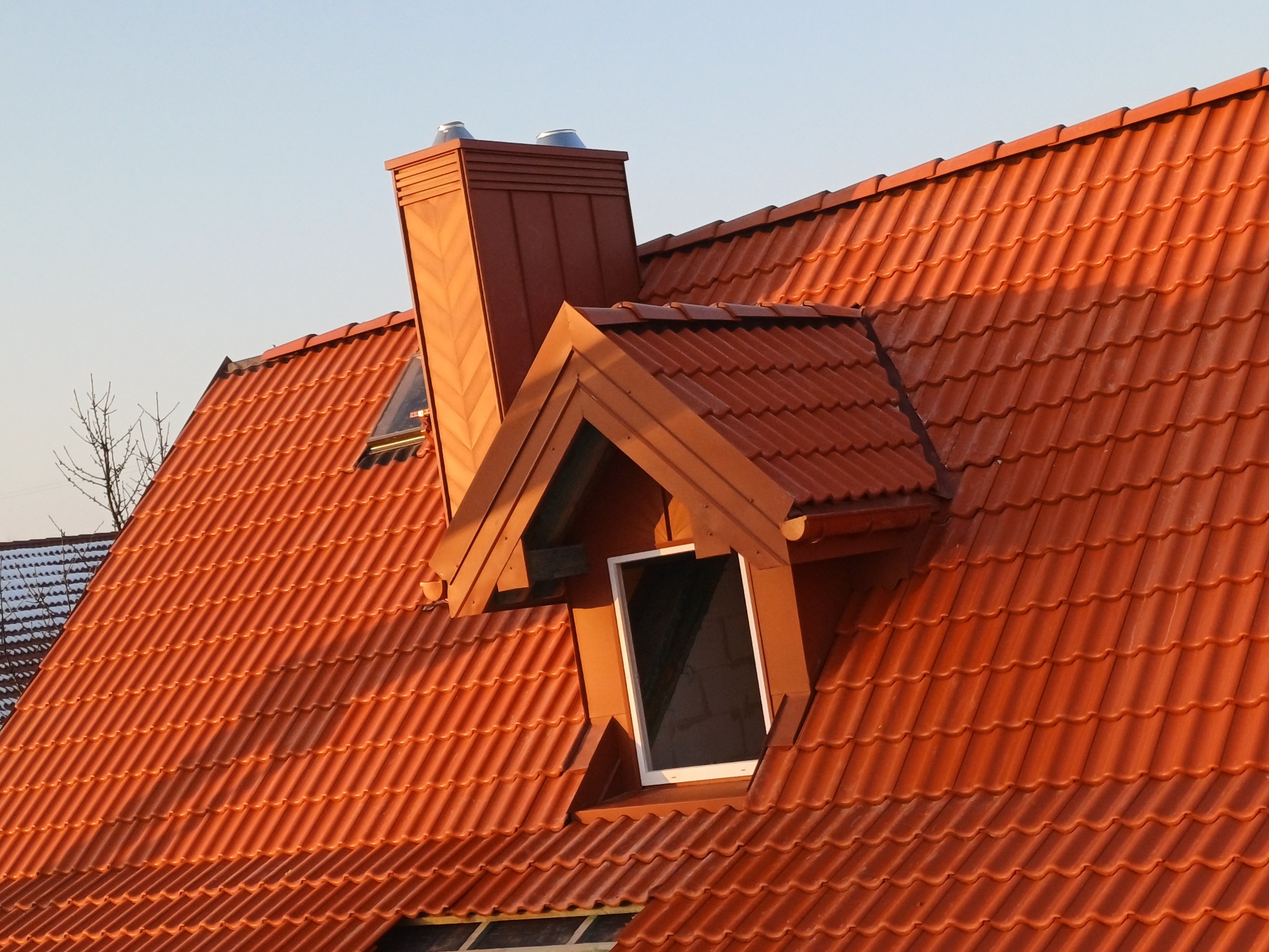 Czerwony dach z dachówki ceramicznej z oknami dachowymi i kominem w ciepłym świetle słonecznym. Widok z góry na nową konstrukcję dachu.