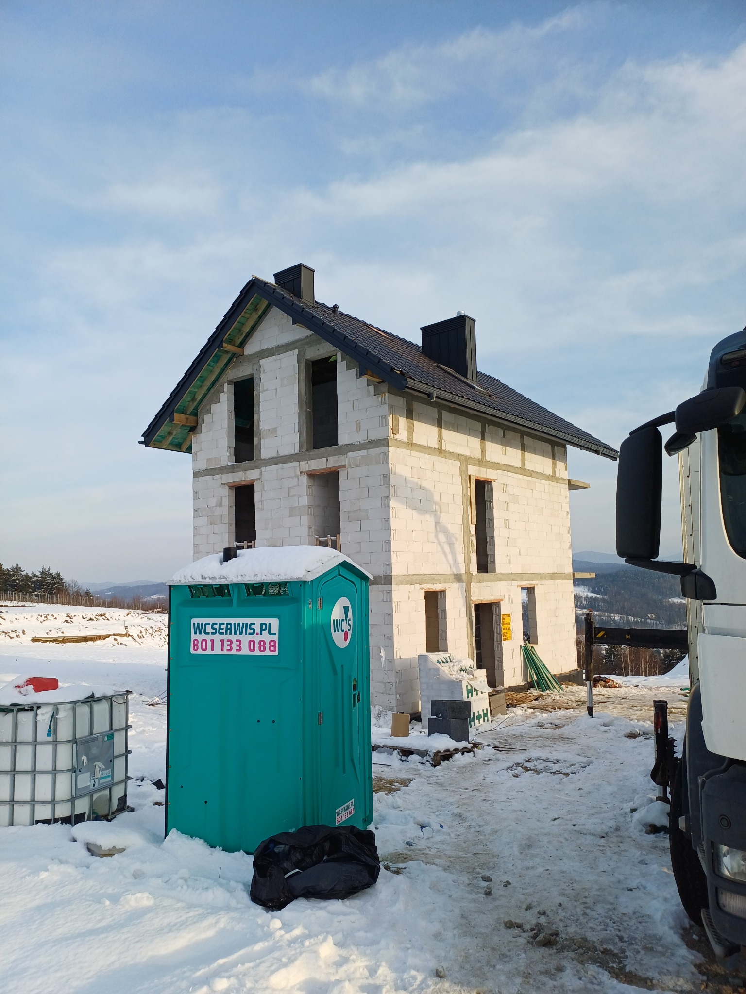 Surowy dom z czarnym dachem w zimowym krajobrazie, widoczna przenośna toaleta i elementy budowlane. Dom w trakcie budowy, z widoczną konstrukcją dachu.