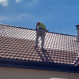 VIPDOMY.PL - Pracownik w jasnej koszulce i szarych spodniach stoi na brązowym dachu z metalowej dachówki, prawdopodobnie podczas jego czyszczenia lub konserwacji, z widocznym kominem zabezpieczonym folią...