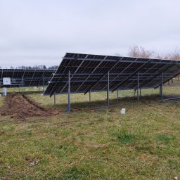 VIPDOMY.PL - Instalacja paneli słonecznych na metalowej konstrukcji w terenie trawiastym, widoczne wykopy pod kable, biały inwerter i pojemnik na wodę.