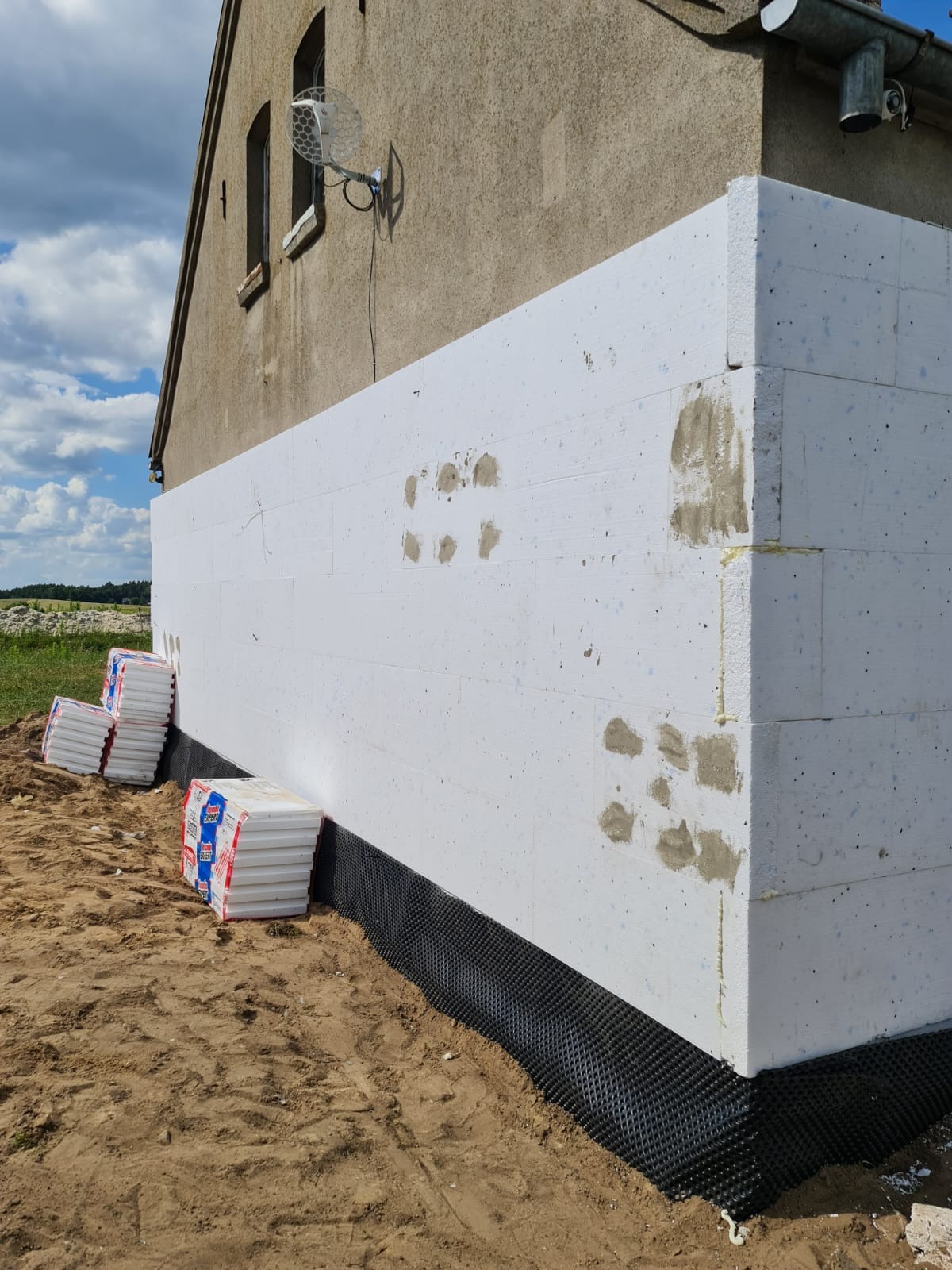 Ocieplanie elewacji budynku styropianem, widoczne pakiety styropianu przy ścianie i fragment elewacji pokryty izolacją, ślady po kleju na styropianie.