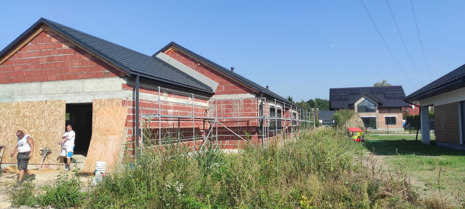 Budowa domu jednorodzinnego w trakcie wznoszenia ścian z czerwonej cegły, widoczne rusztowania i pracownicy, w tle inne domy z ciemnymi dachami.