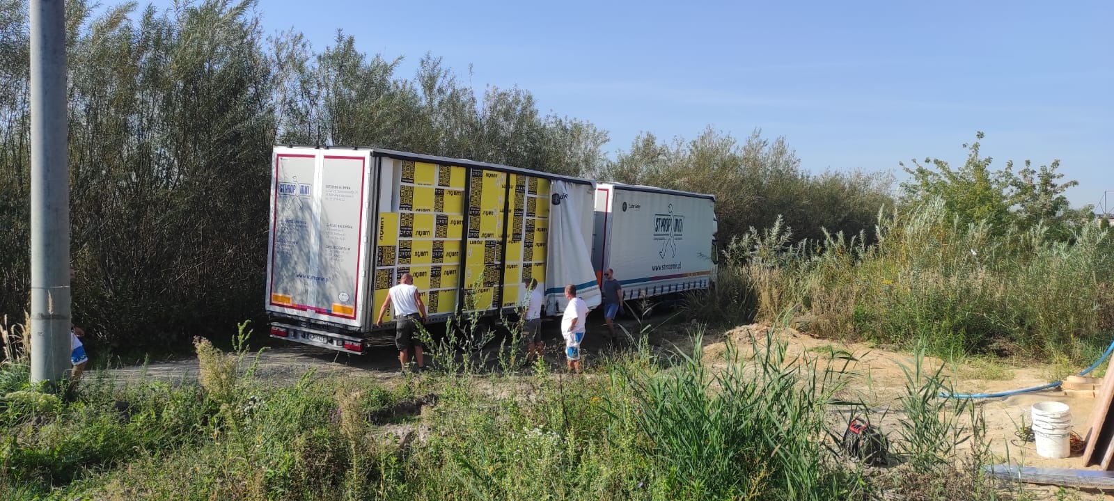Rozładunek transportu płyt styropianowych z ciężarówki na zewnątrz budynku, widoczni trzej mężczyźni w trakcie pracy.
