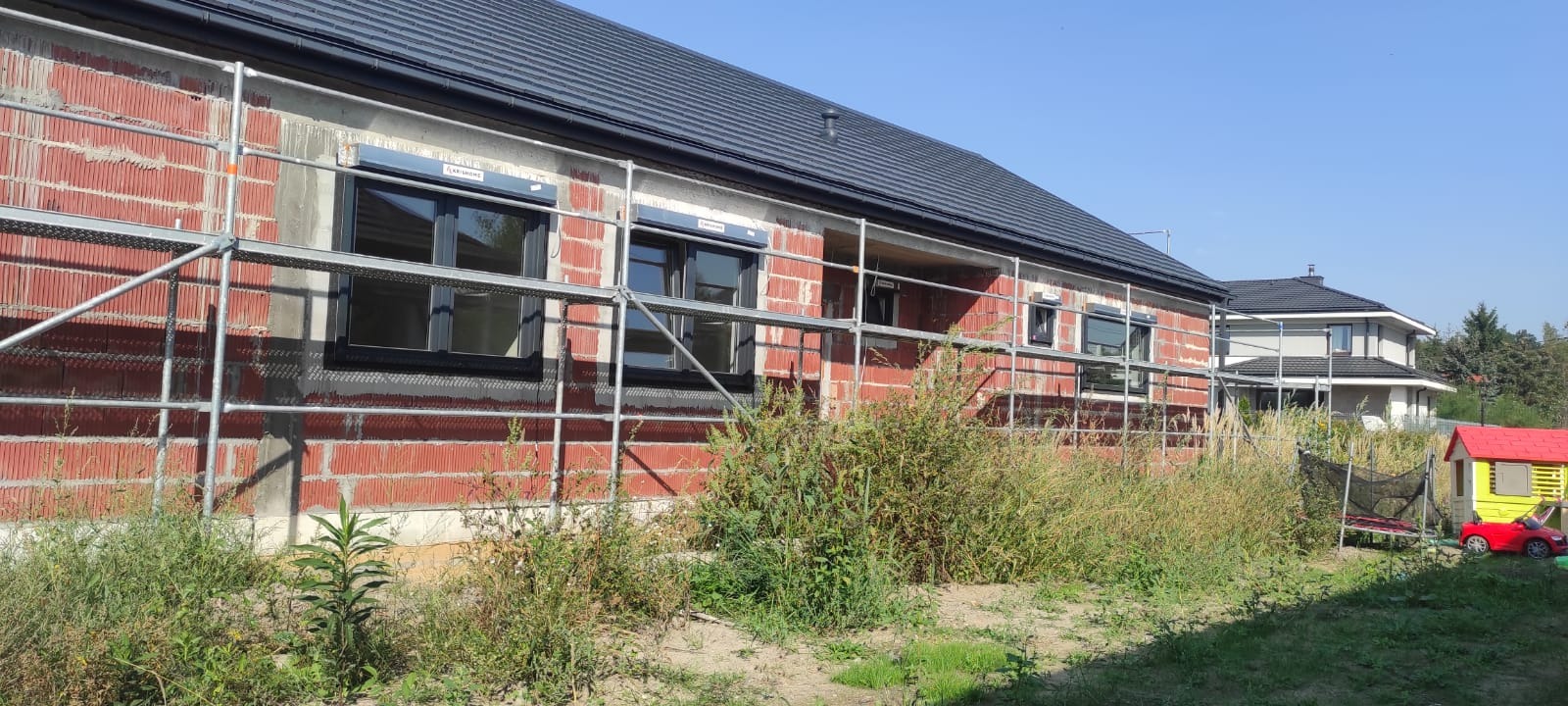 Dom w trakcie budowy, widoczne rusztowania przy elewacji z czerwonej cegły, czarne okna z roletami zewnętrznymi, dach pokryty ciemną dachówką, bujna roślinność w ogrodzie, w tle drugi dom i zabawki...