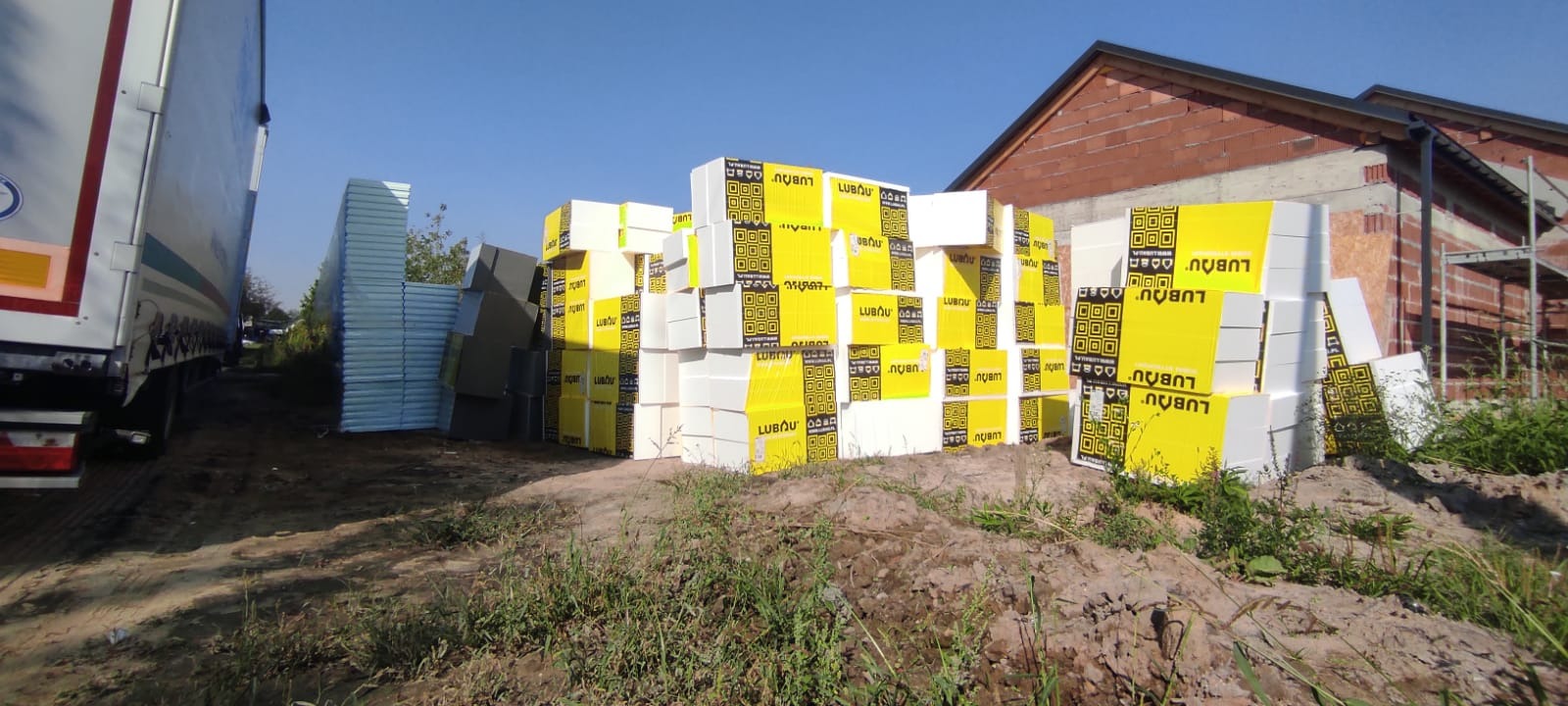 Stosy styropianu marki Lubau w żółtych opakowaniach na placu budowy, obok zaparkowana naczepa ciężarówki, w tle surowa bryła budynku z czerwonej cegły i rusztowania.