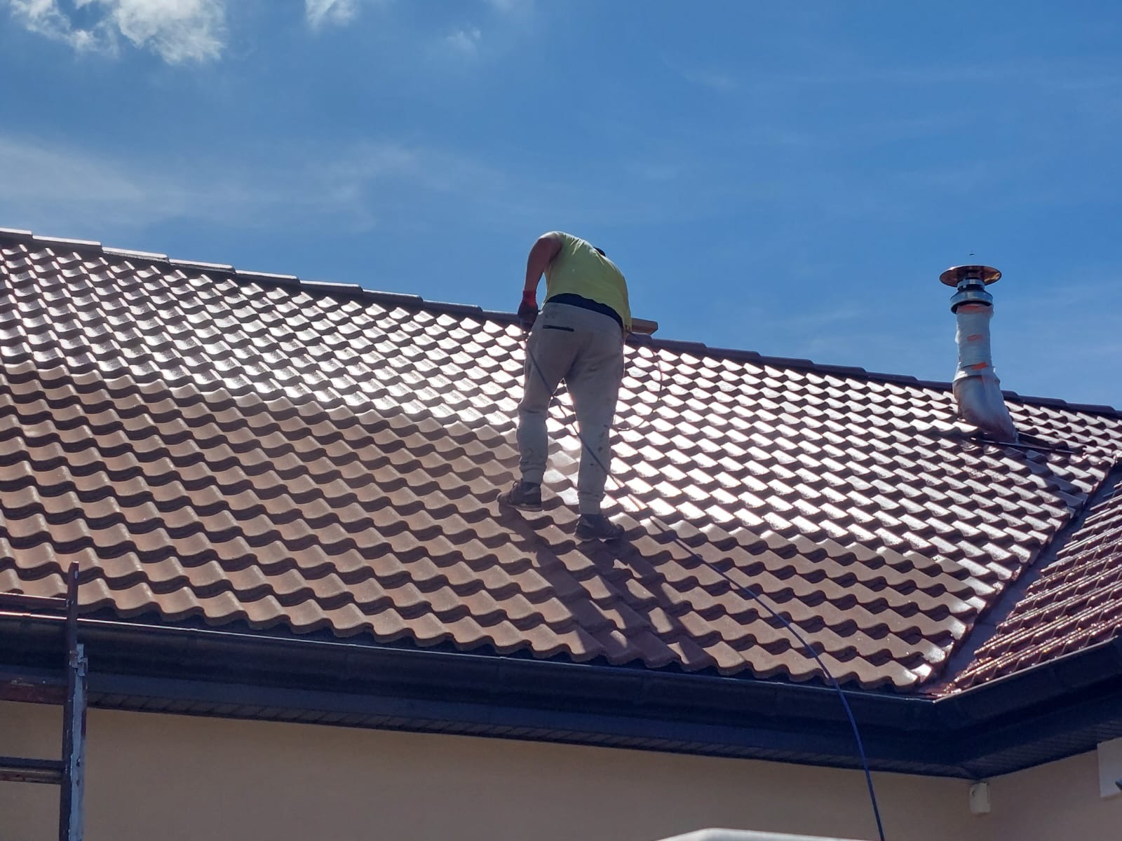 Pracownik w jasnej koszulce i szarych spodniach stoi na brązowym dachu z metalowej dachówki, prawdopodobnie podczas jego czyszczenia lub konserwacji, z widocznym kominem zabezpieczonym folią...