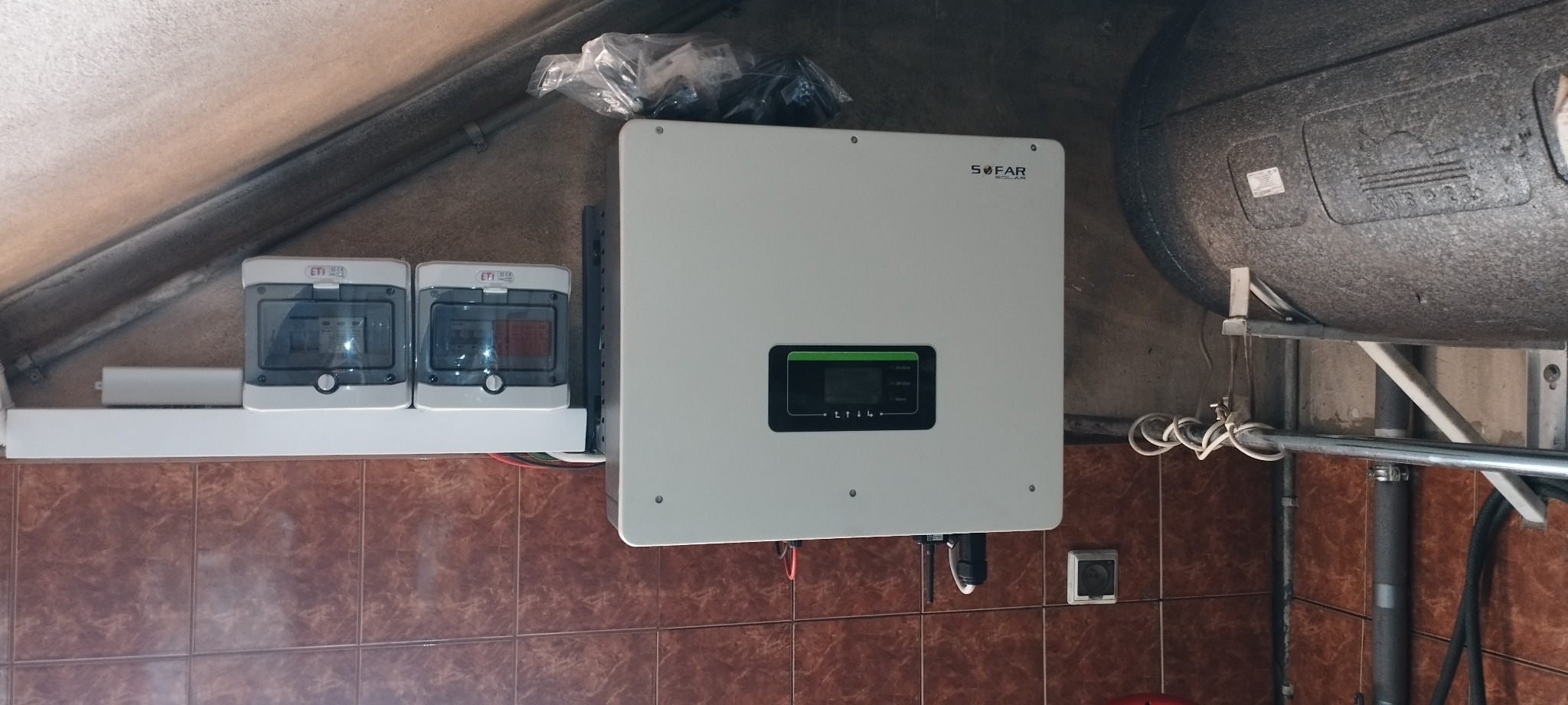 Zamontowany biały inwerter solarny marki Sofar Solar z podłączonymi przewodami, widoczne zabezpieczenia elektryczne ETI w obudowach oraz zasobnik ciepłej wody użytkowej w kotłowni z brązowymi...