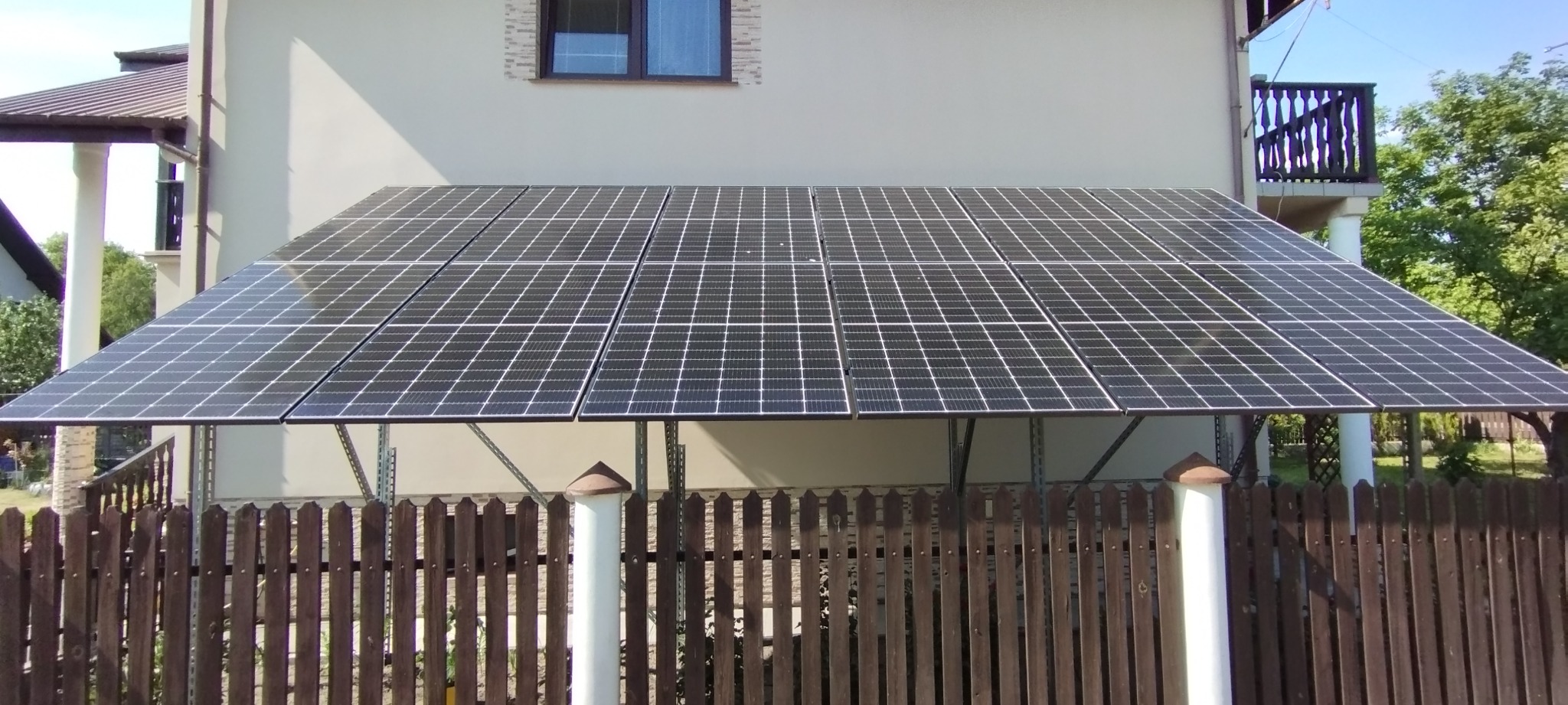 Panele fotowoltaiczne zamontowane jako zadaszenie nad posesją, widoczne zza drewnianego płotu.