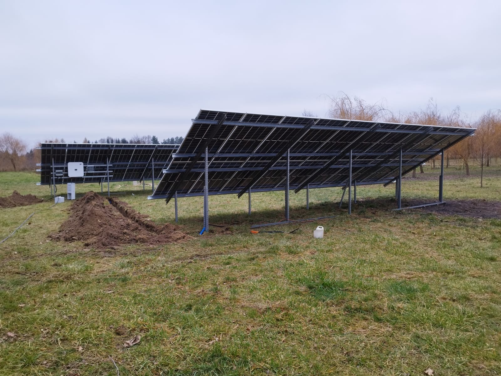 Instalacja paneli słonecznych na metalowej konstrukcji w terenie trawiastym, widoczne wykopy pod kable, biały inwerter i pojemnik na wodę.