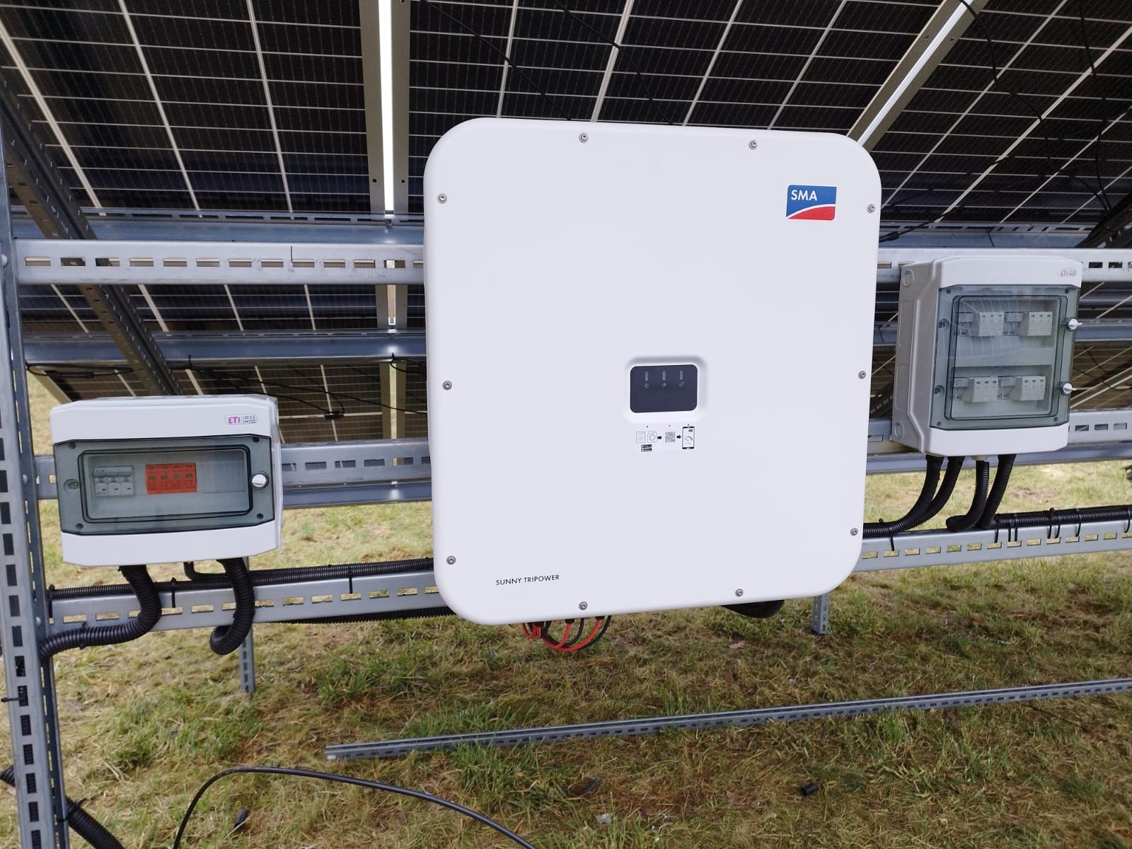 Biały inwerter SMA Sunny Tripower i skrzynki elektryczne zamontowane na metalowej konstrukcji pod panelami słonecznymi na trawiastym terenie.