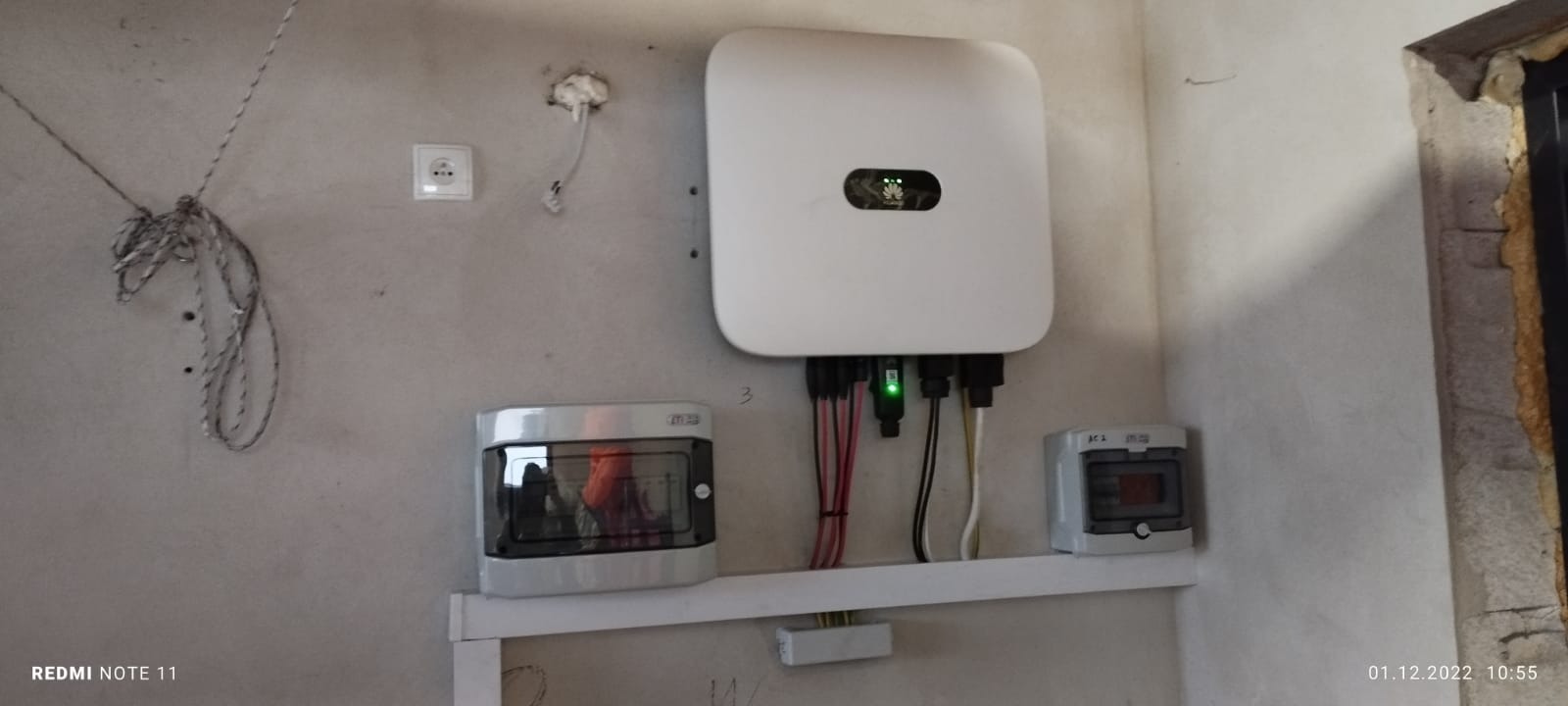 Biały inwerter Huawei zamontowany na szarej ścianie z widocznymi przewodami, puszkami elektrycznymi i gniazdkiem. Ściana jest surowa, w trakcie wykończenia.