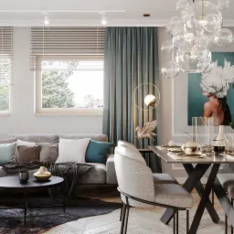 Jasny salon z szarą sofą narożną, okrągłym stolikiem kawowym, jadalnią z czarnym stołem na krzyżakowych nogach i dekoracyjną lampą wiszącą z przezroczystych kul. Na ścianie obraz z motywem...