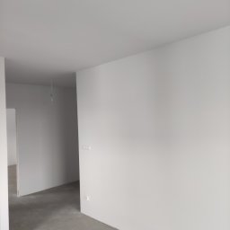 Molart - Wnętrze po remoncie: białe ściany, szara podłoga, widoczne gniazdka elektryczne. Minimalistyczny styl wykończenia, surowy beton na podłodze. W tle widoczne kolejne pomieszczenia.