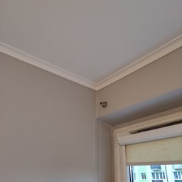 Molart - Pokój po odświeżeniu: ściany pomalowane na szaro, biały sufit z dekoracyjną listwą przy suficie, okno z roletą w tle. Widoczny fragment ściany i sufitu.